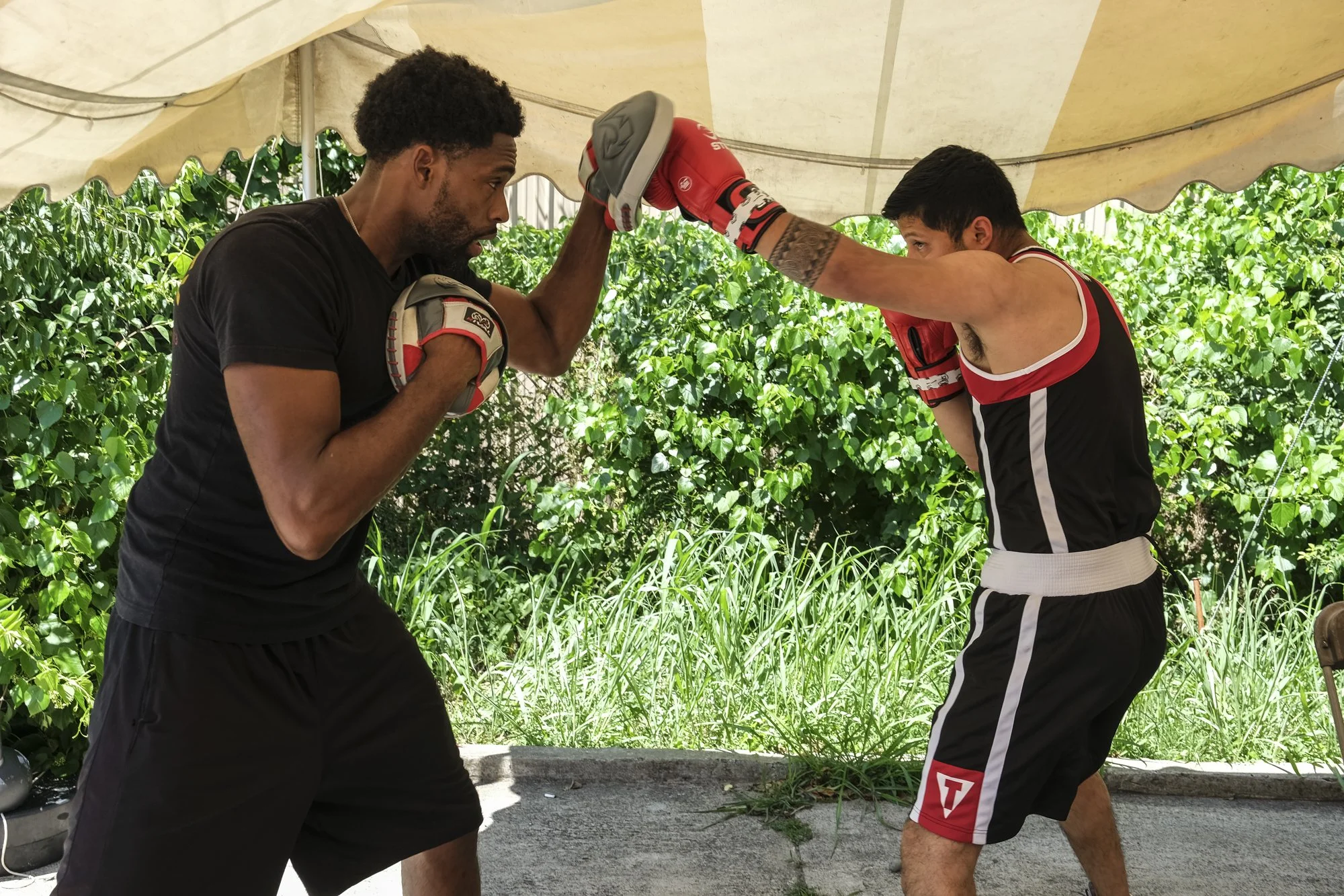 Two men practicing boxing outdoors under a tent, with one man wearing black shirts and the other wearing black and red boxing gear, during the daytime.