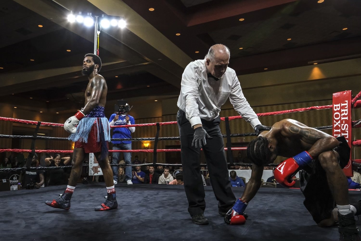 A boxing match is taking place inside a ring, with one boxer standing confidently and the other crouched on the ground, possibly injured or exhausted. A referee is inspecting the downed boxer, and a cameraman is capturing the scene. Audience members 