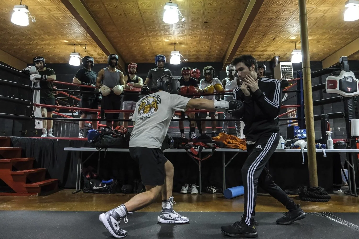 A boxing training session with a person practicing punches on a coach inside a boxing gym, with the coach blocking the punches. Several people, all wearing boxing gear, watch from the background, standing behind a boxing ring.