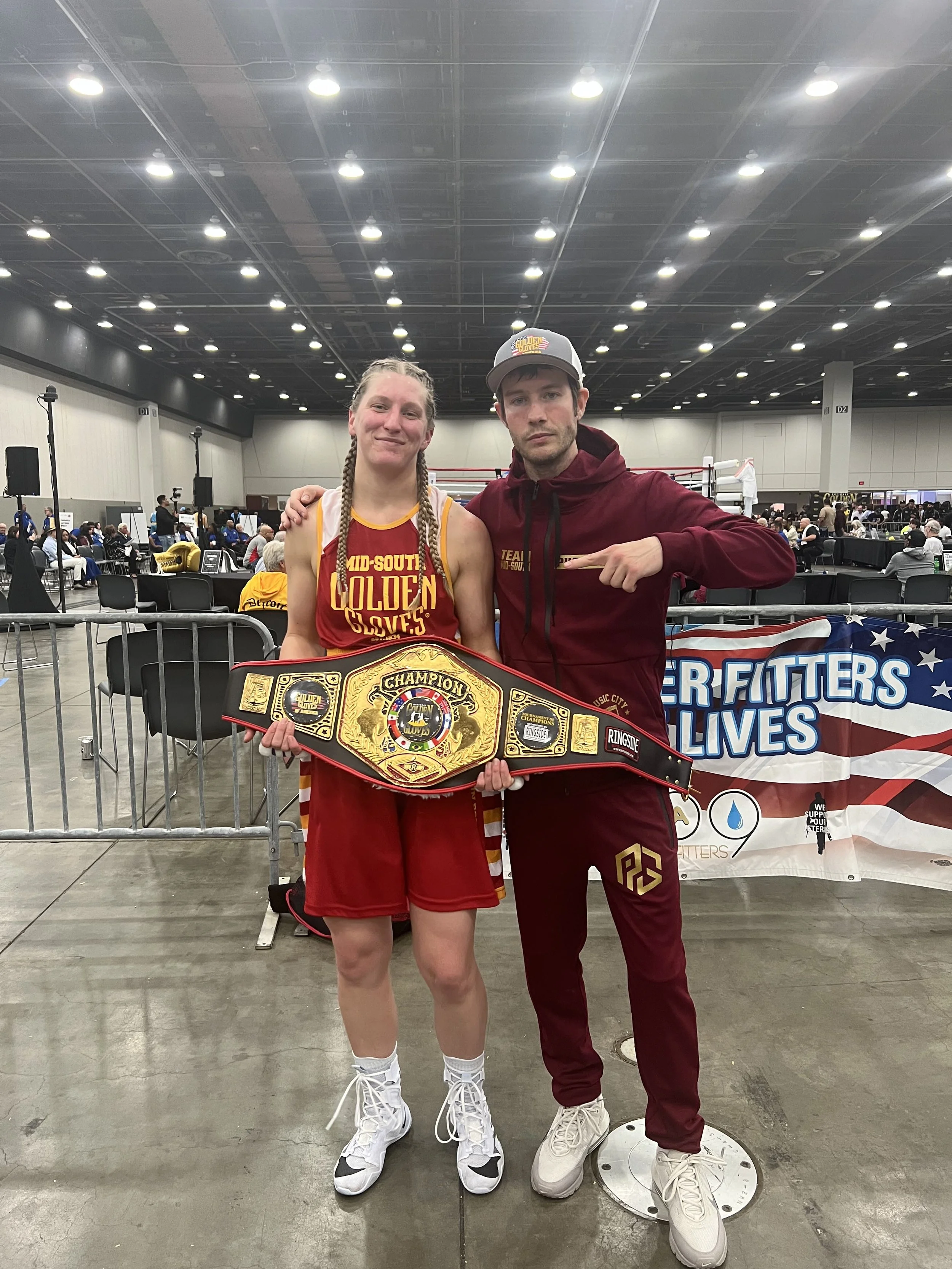 A female boxer in red and yellow athletic gear holding a championship belt, standing next to a male coach or trainer in maroon athletic wear inside a boxing or sports arena.