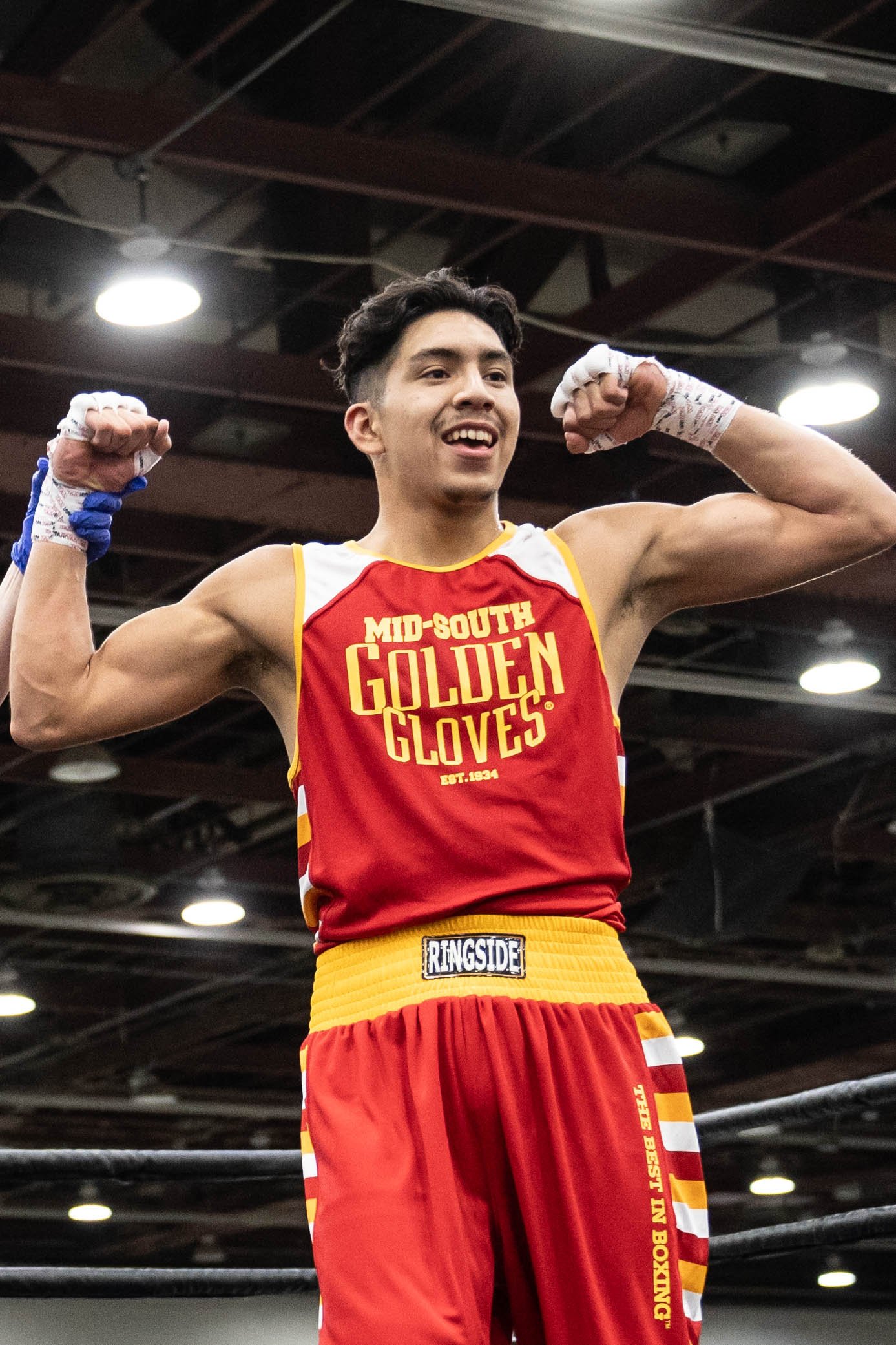 A young male boxer in red and yellow shorts and matching red tank top with 'Mid-South Golden Gloves' printed on it, flexing his biceps inside a boxing gym.