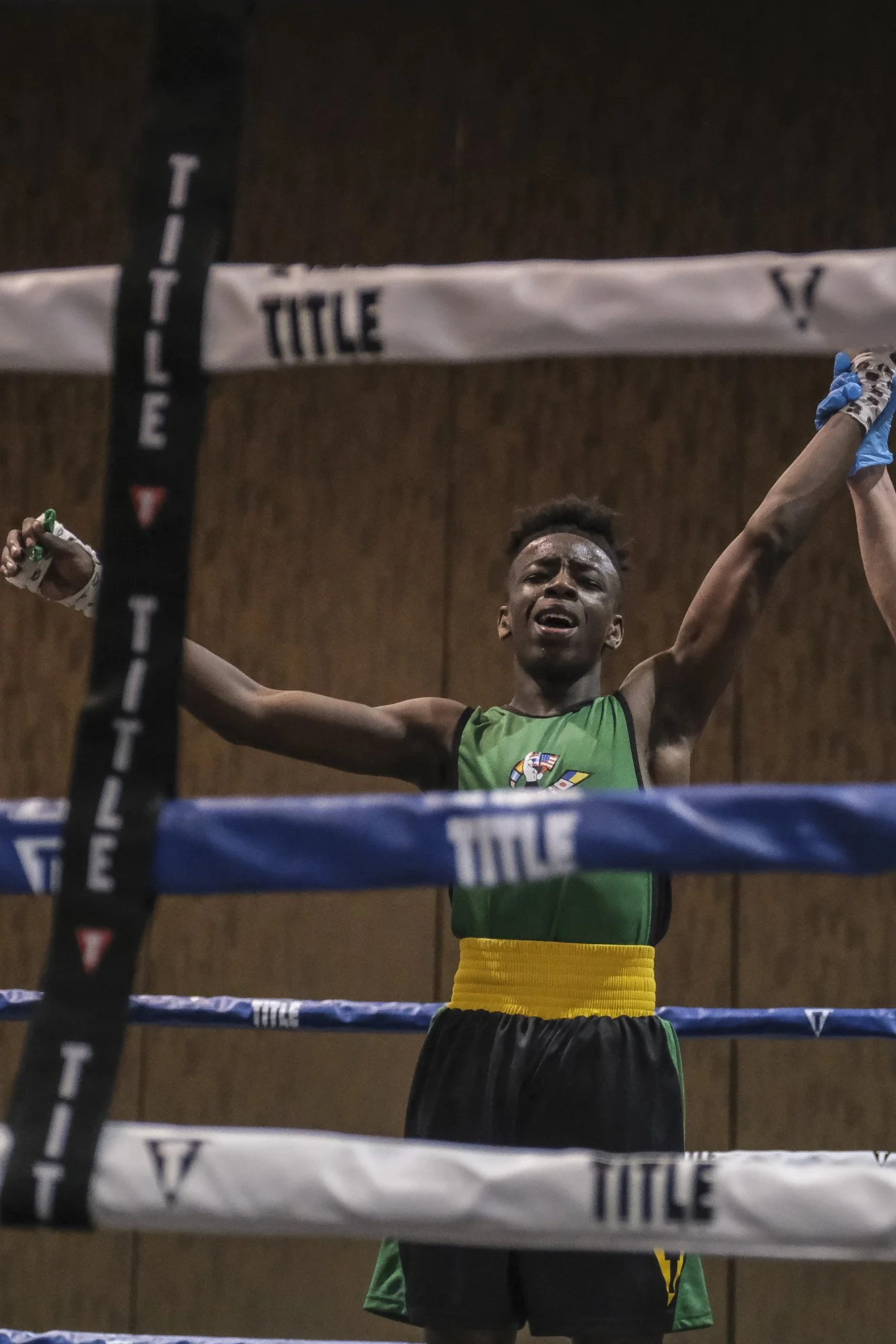 A young male boxer in green shorts and a green and black tank top with a yellow waistband, standing inside a boxing ring with his arms raised and a look of triumph on his face.