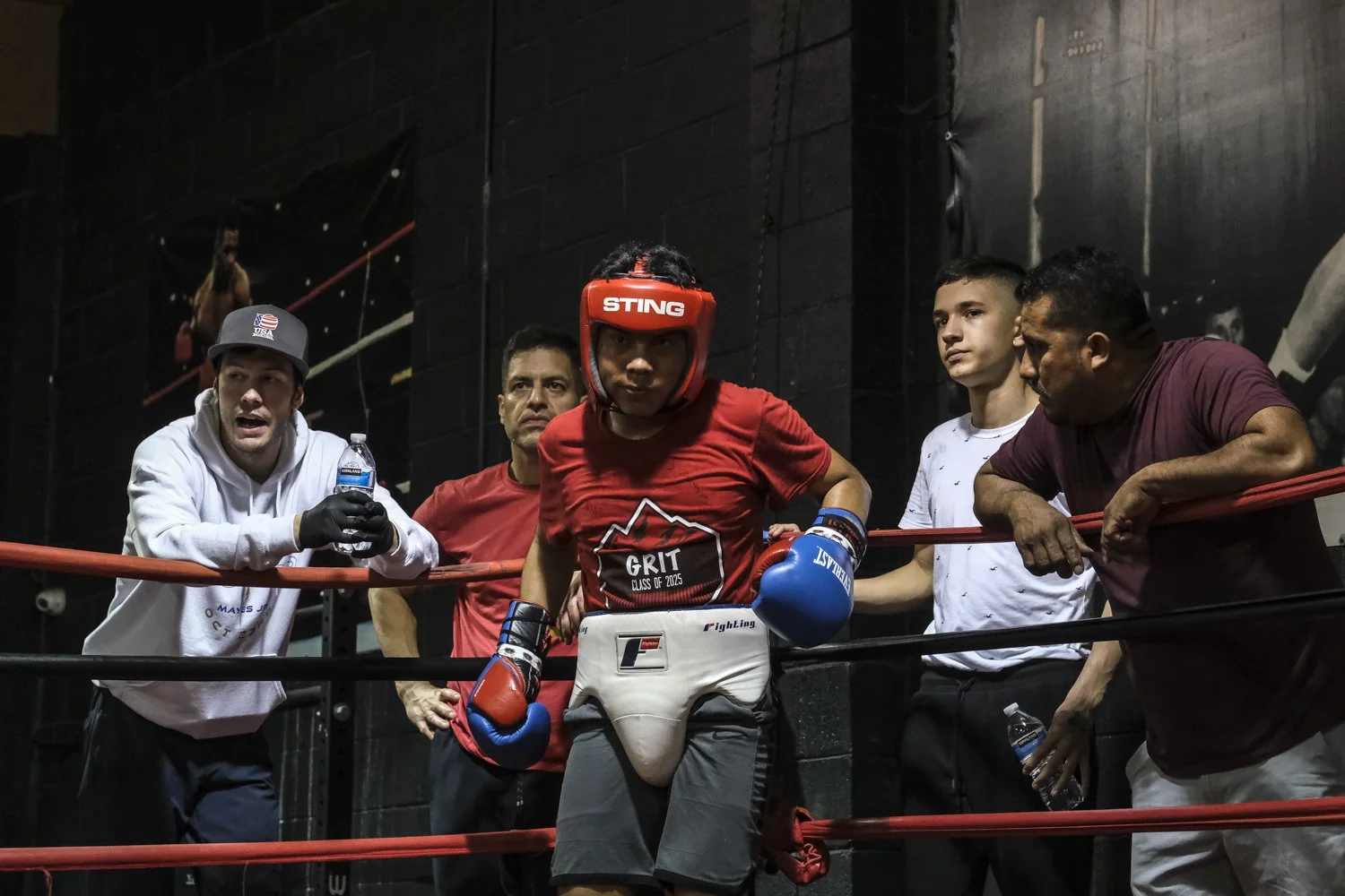 A young female boxer in red gear standing in a boxing ring with four men around her, one holding a water bottle, in a gym or training facility.