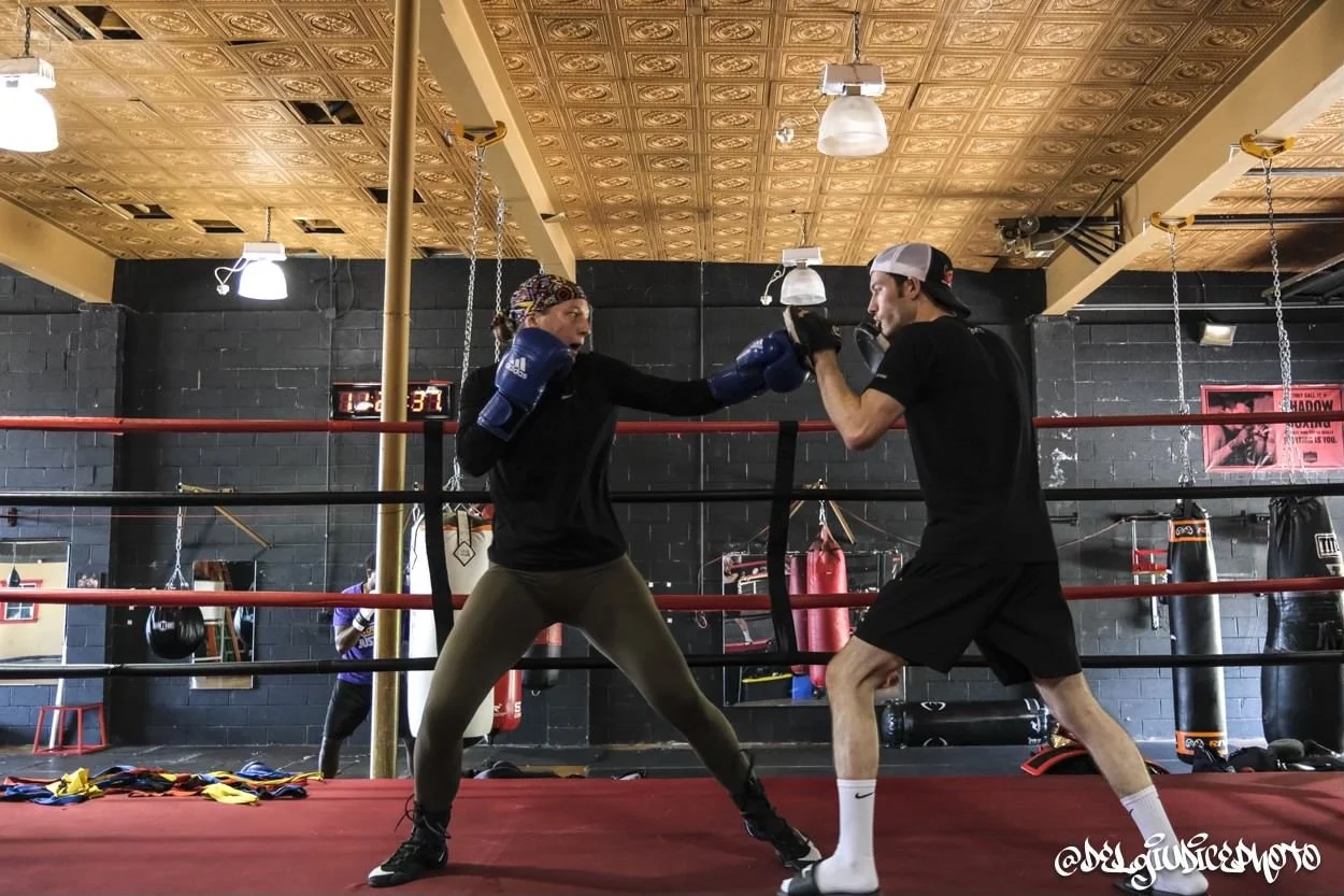 A woman in boxing gloves and athletic gear sparring with a man wearing gloves and a backwards cap inside a boxing gym with black brick walls, punching each other in a boxing ring.