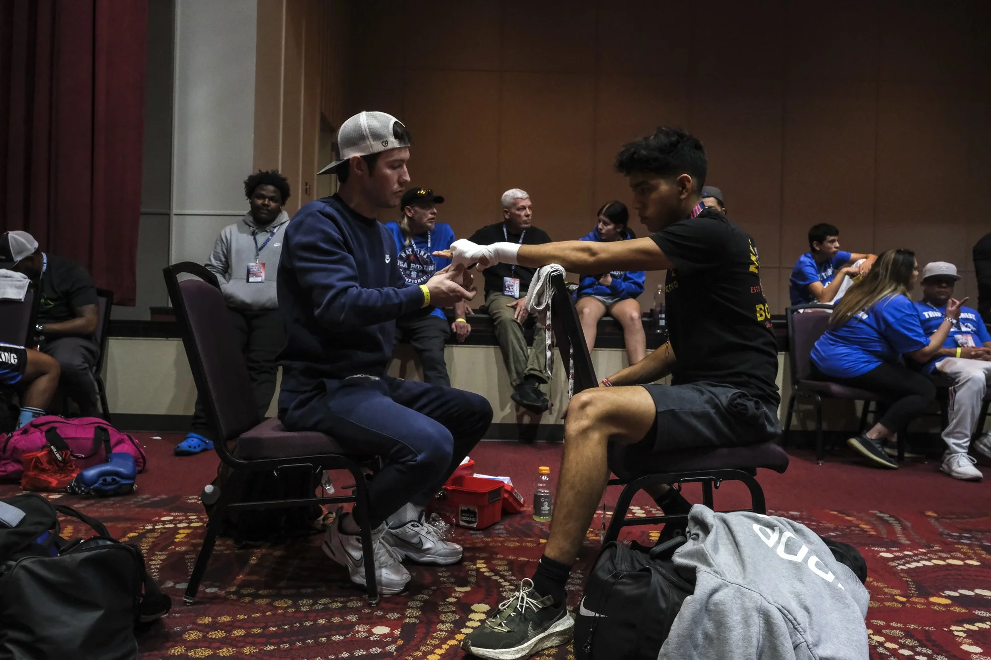 Two young male boxers are sitting face-to-face in a sports arena, wrapping hands, with a group of people watching in the background.