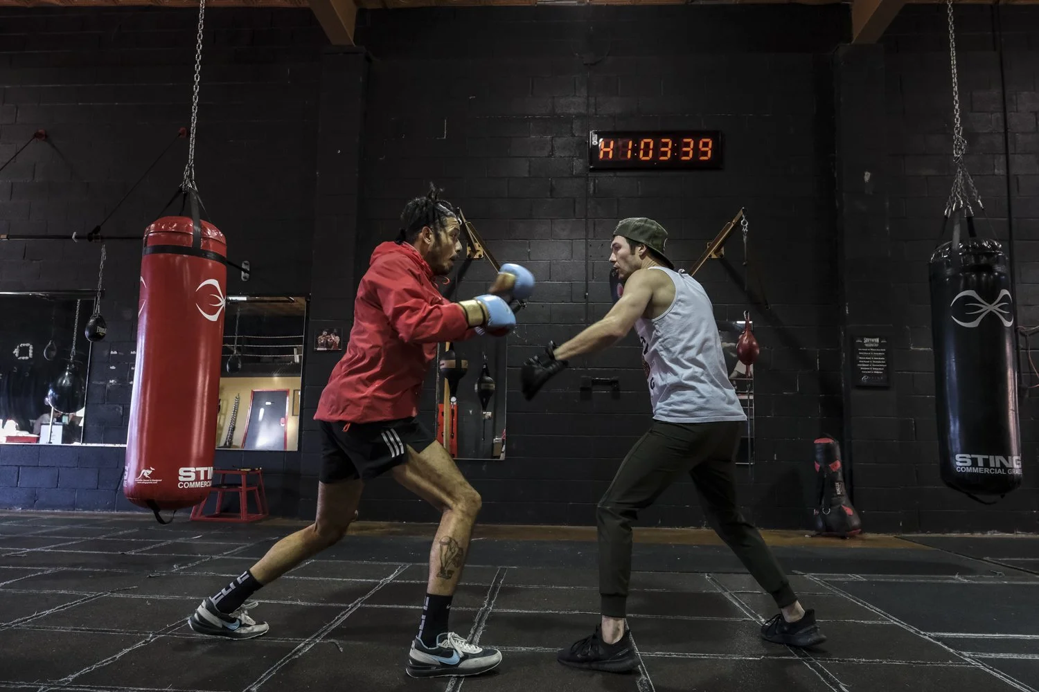 Two men boxing in a gym. One man in a red hoodie and black shorts with boxing gloves. The other man in a gray tank top, black pants, and a cap, also with boxing gloves. They are sparring with a black punching bag on the right. A timer on the wall rea