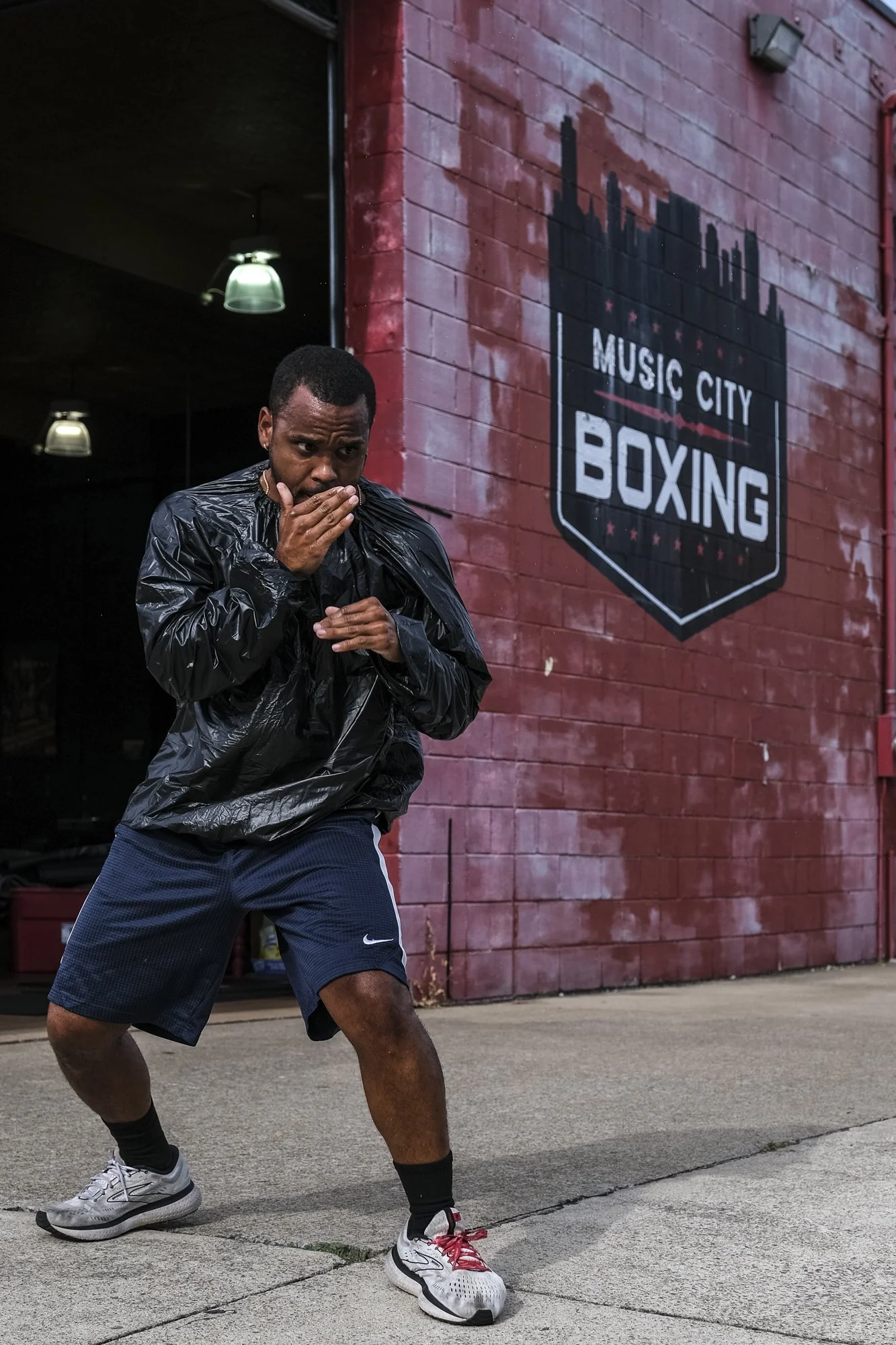 A man wearing a black rain jacket, navy shorts, and sneakers practices boxing outside a gym with a sign that reads 'Music City Boxing' on a red brick wall.