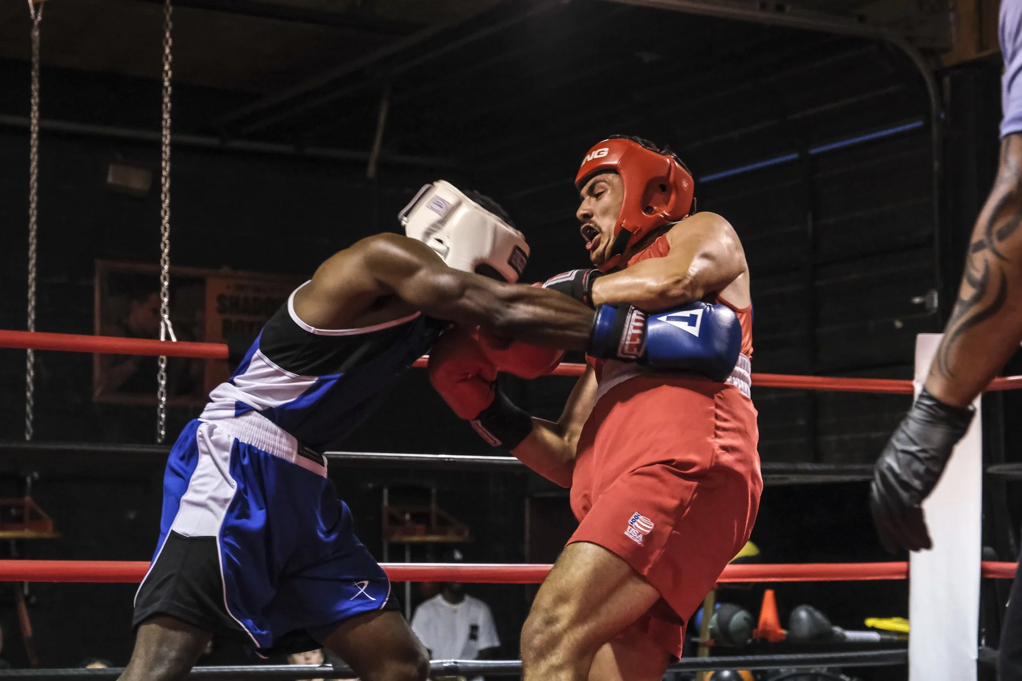 Two male boxers in boxing gloves and protective headgear engaged in a fight inside a boxing ring, one wearing black and white shorts and the other in red shorts with a U.S. flag patch, under low lighting.