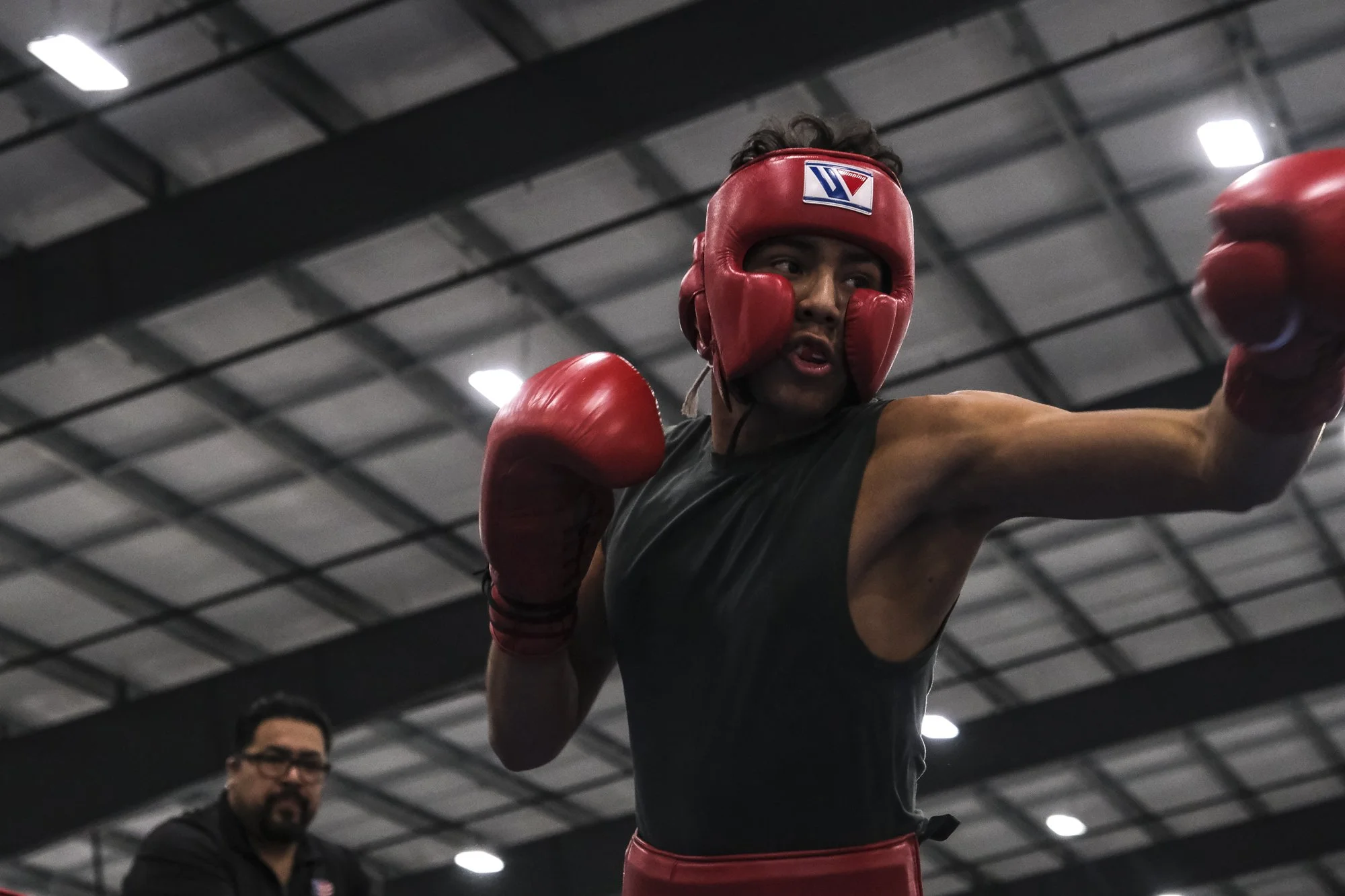 A man wearing red boxing gloves, a red headgear, and a black sleeveless shirt practicing boxing in a gym.