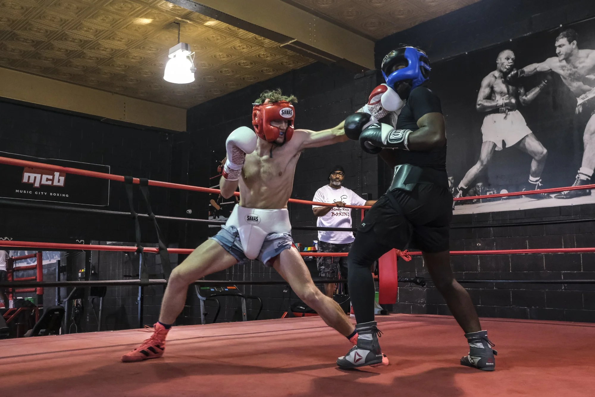 Two boxers sparring in a boxing ring, one in red gear and the other in blue gear, with a coach in the background watching.