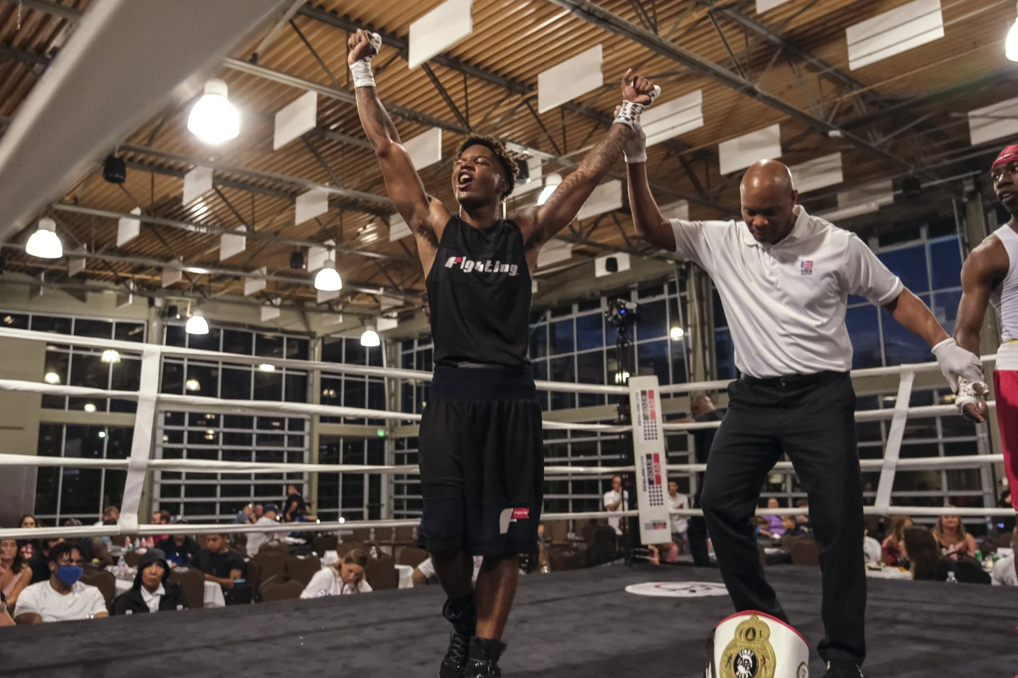 A victorious boxer with arms raised in victory inside a boxing ring, with a referee holding up his arm. The crowd watches from the background in an indoor sports arena.