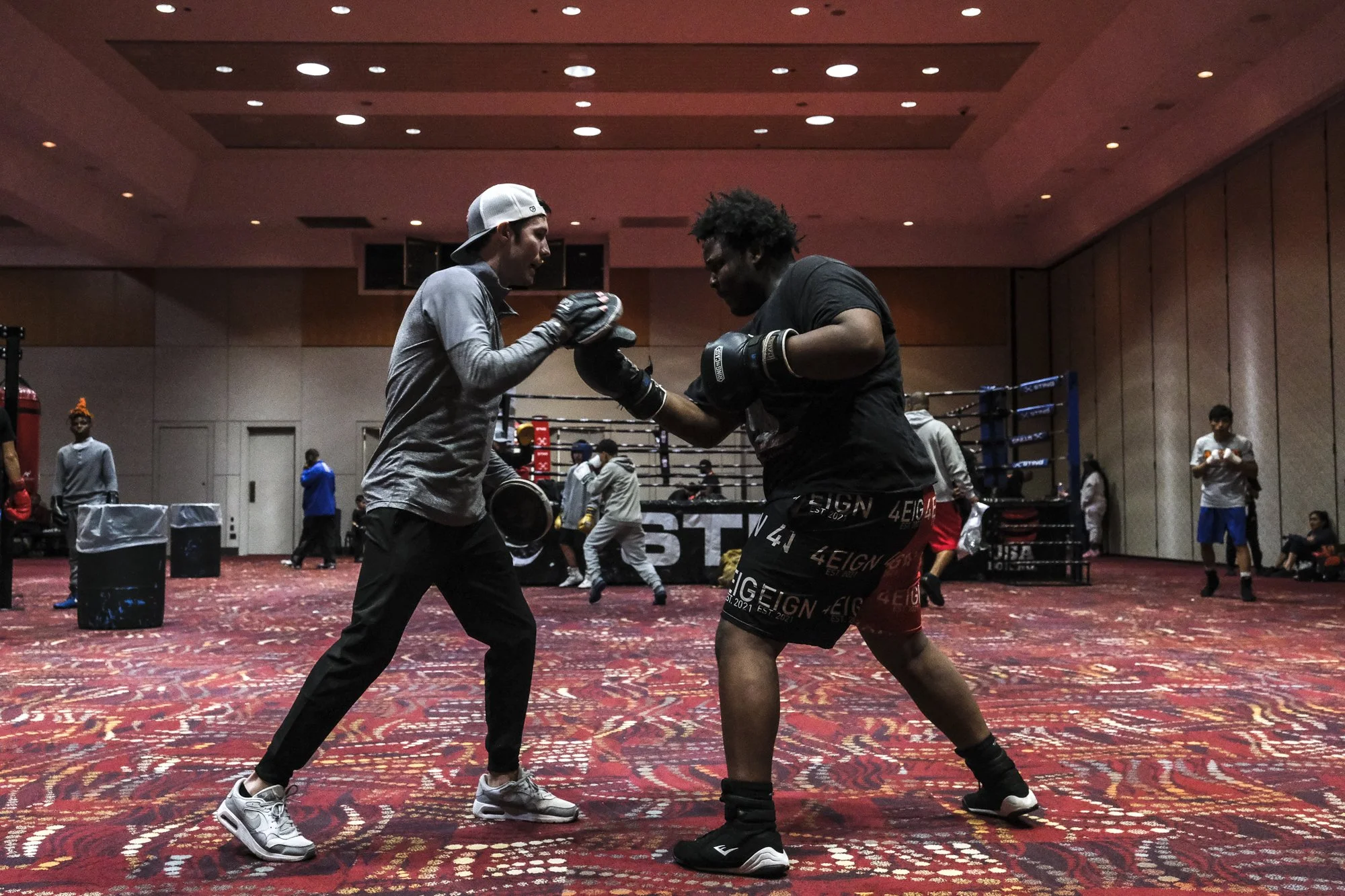 Two men boxing in a training gym with a boxing ring in the background and other people practicing or observing.