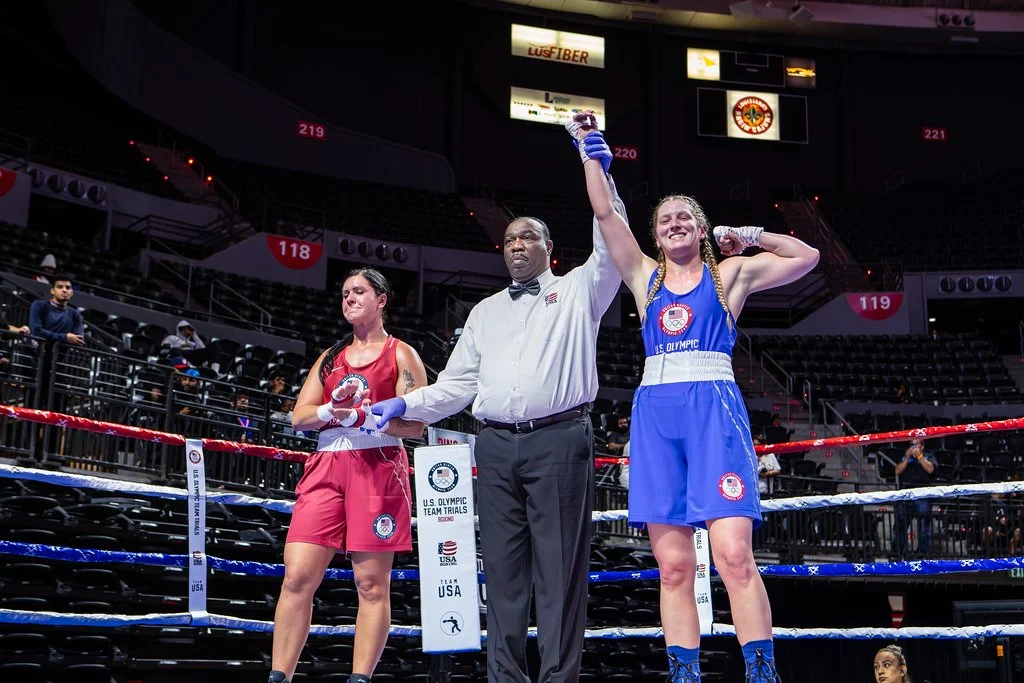 A boxing match ends with the victorious female boxer in blue raising her right hand by the referee, while her opponent in red looks down. They are inside a boxing ring with empty seats and a few spectators in the background.