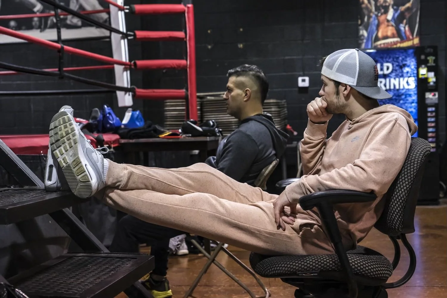 Two men sitting in a gym, one with legs stretched out on a desk, wearing a beige hoodie and cap backward, the other with a short hairstyle, wearing dark workout clothes. Gym equipment and a vending machine are visible in the background.