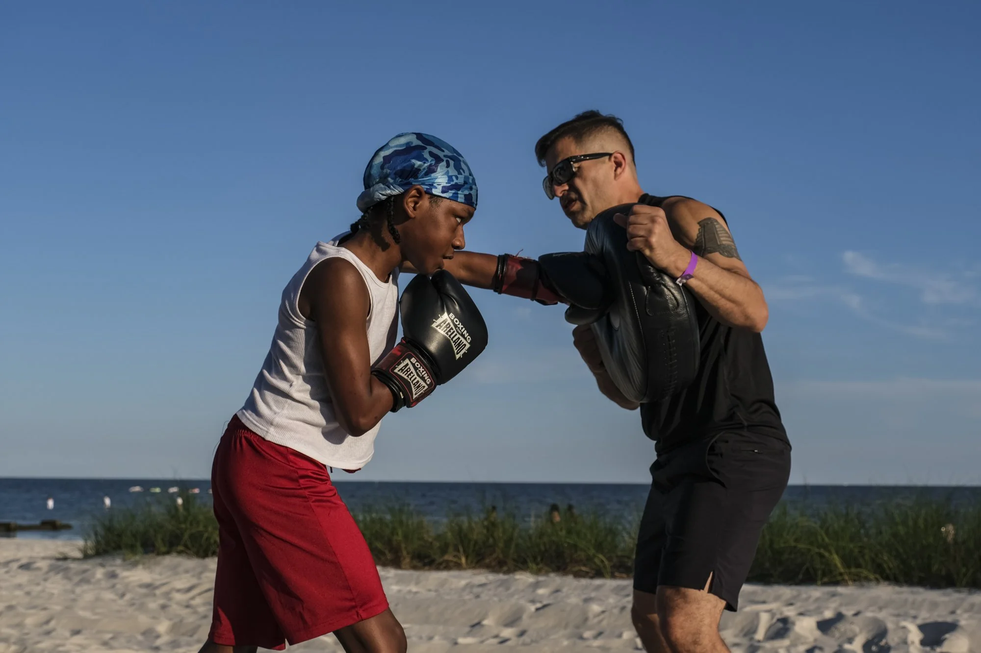 A young woman in boxing gloves and sleeveless shirt practicing boxing with a man carrying boxing pads on a beach with sand, grass, and ocean in the background under blue sky.
