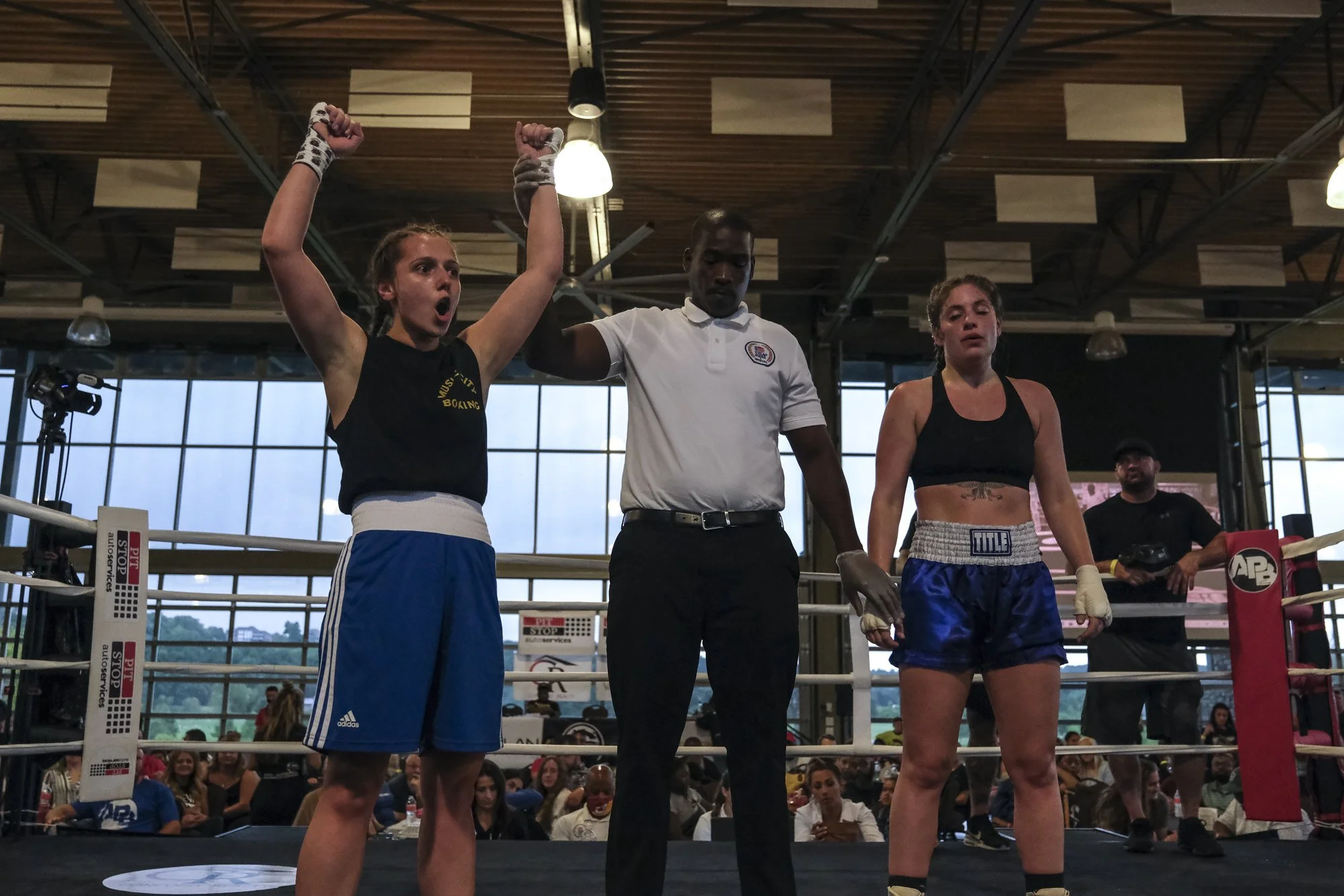Two female boxers in a boxing ring, one with raised fists and the referee holding her arm up, the other with a sad expression and gloves down. The referee is a man in a white shirt. There are spectators in the background.