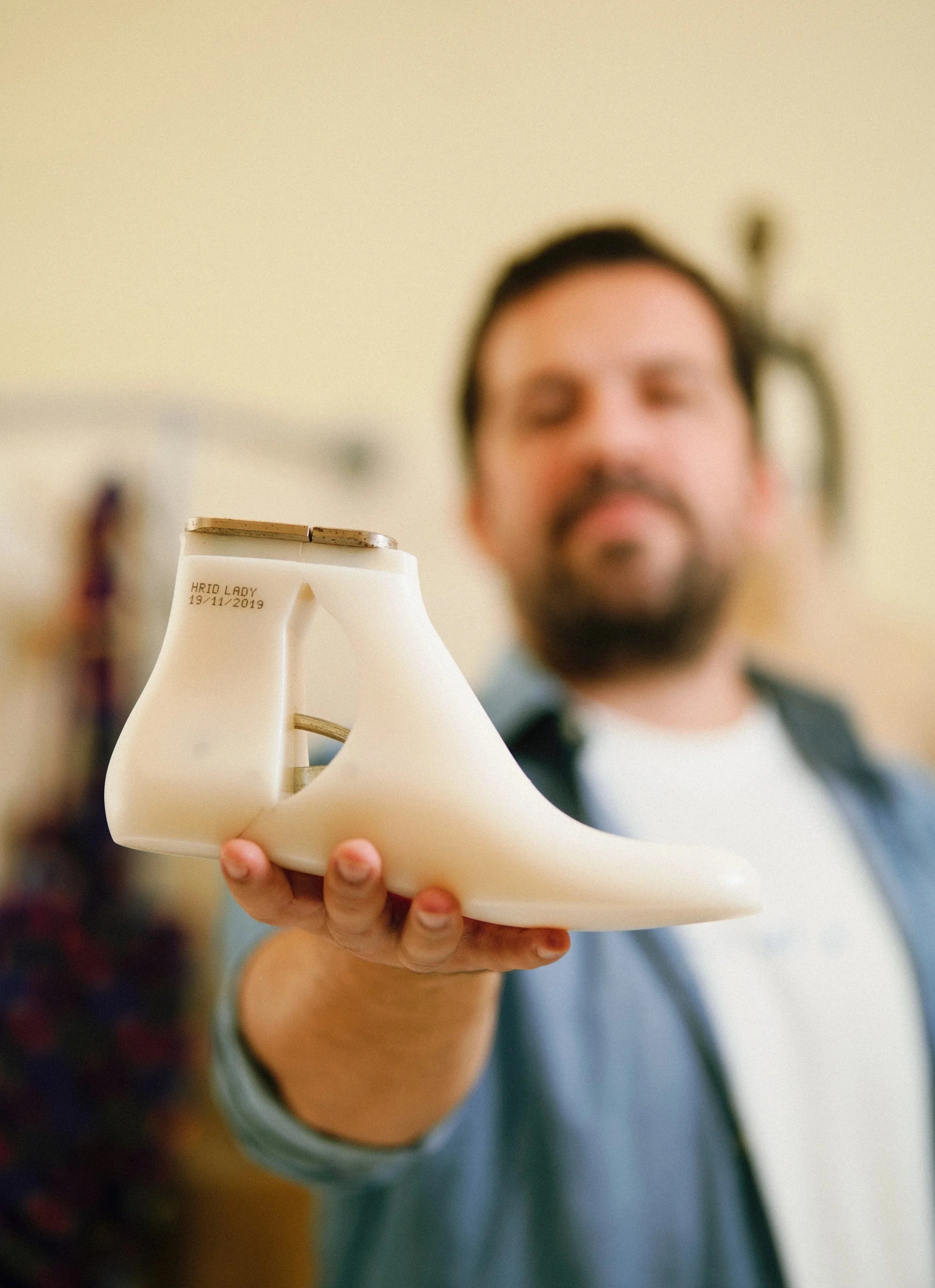 A man is holding a small, off-white, high-heeled shoe-shaped object towards the camera. The shoe has a plaque with the inscription 'HRID LADY 19/11/2019'. The man has a beard and is wearing a shirt.