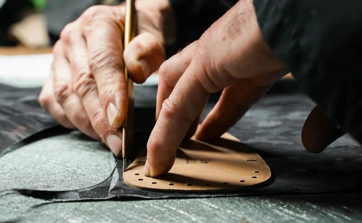 Close-up of a person's hands working on cutting or shaping a piece of tan leather using a craft tool, with a sheet of paper and cutting mat in the background.