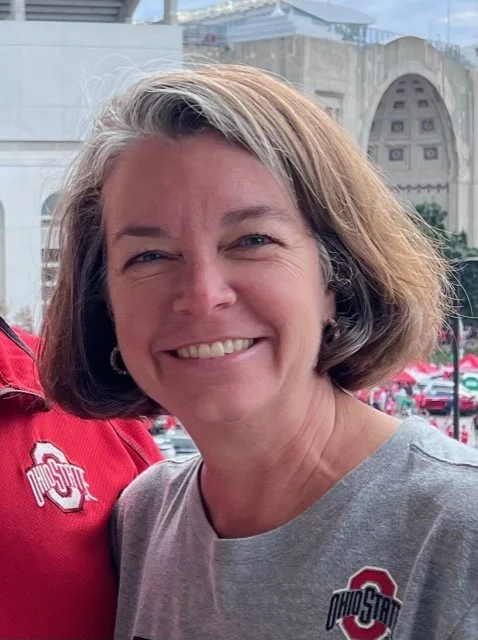 A woman with shoulder-length brown hair smiling outdoors, wearing a gray Ohio State t-shirt with an Ohio State logo, and standing next to someone in a red Ohio State shirt. In the background, there are buildings and a parking lot.