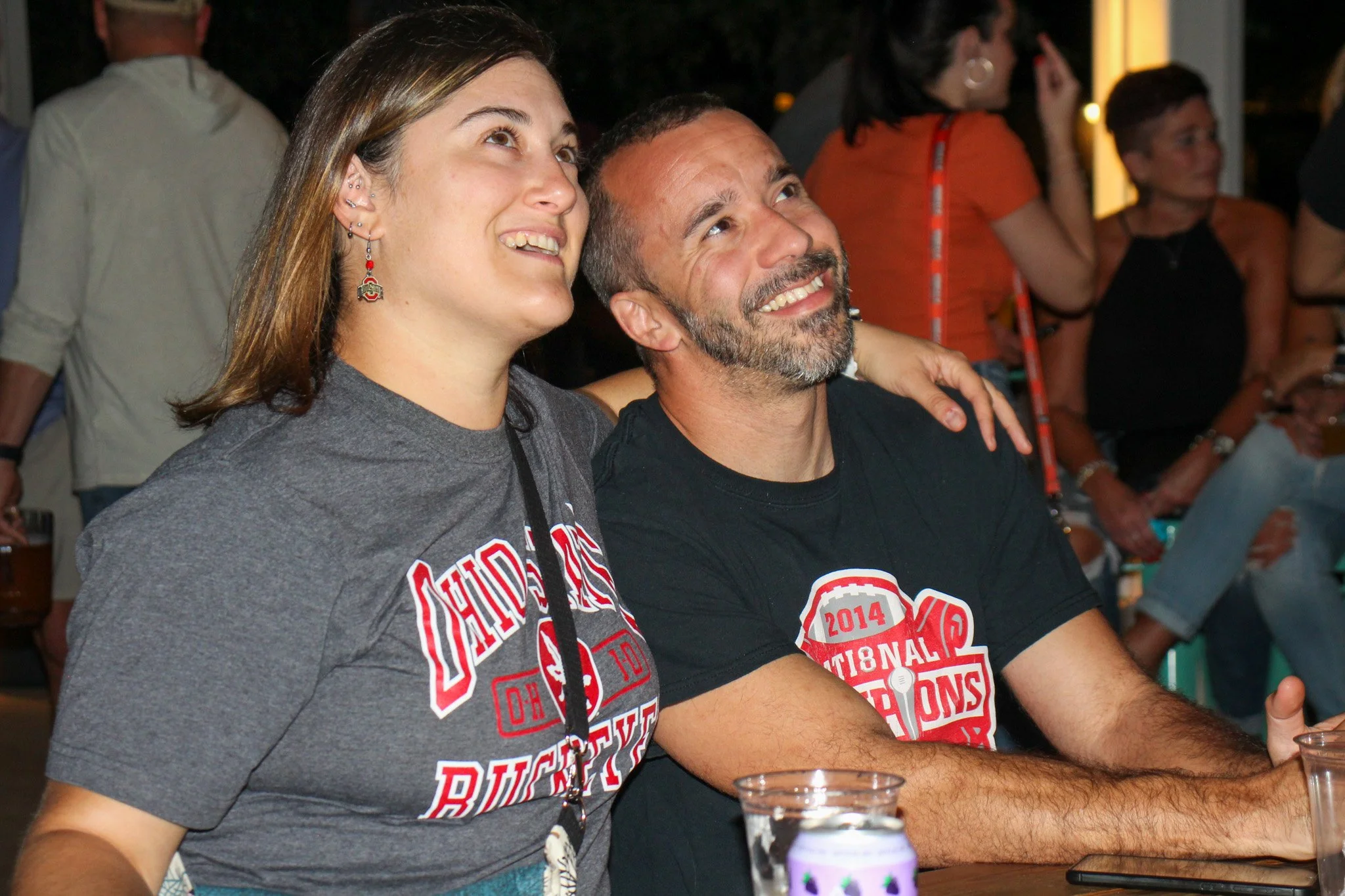 A young woman and an older man sit close together, smiling and looking upward, at a social gathering. They are wearing black and gray shirts with Ohio State Buckeyes logos and the year 2014 SiN at the center. The background shows other people standin