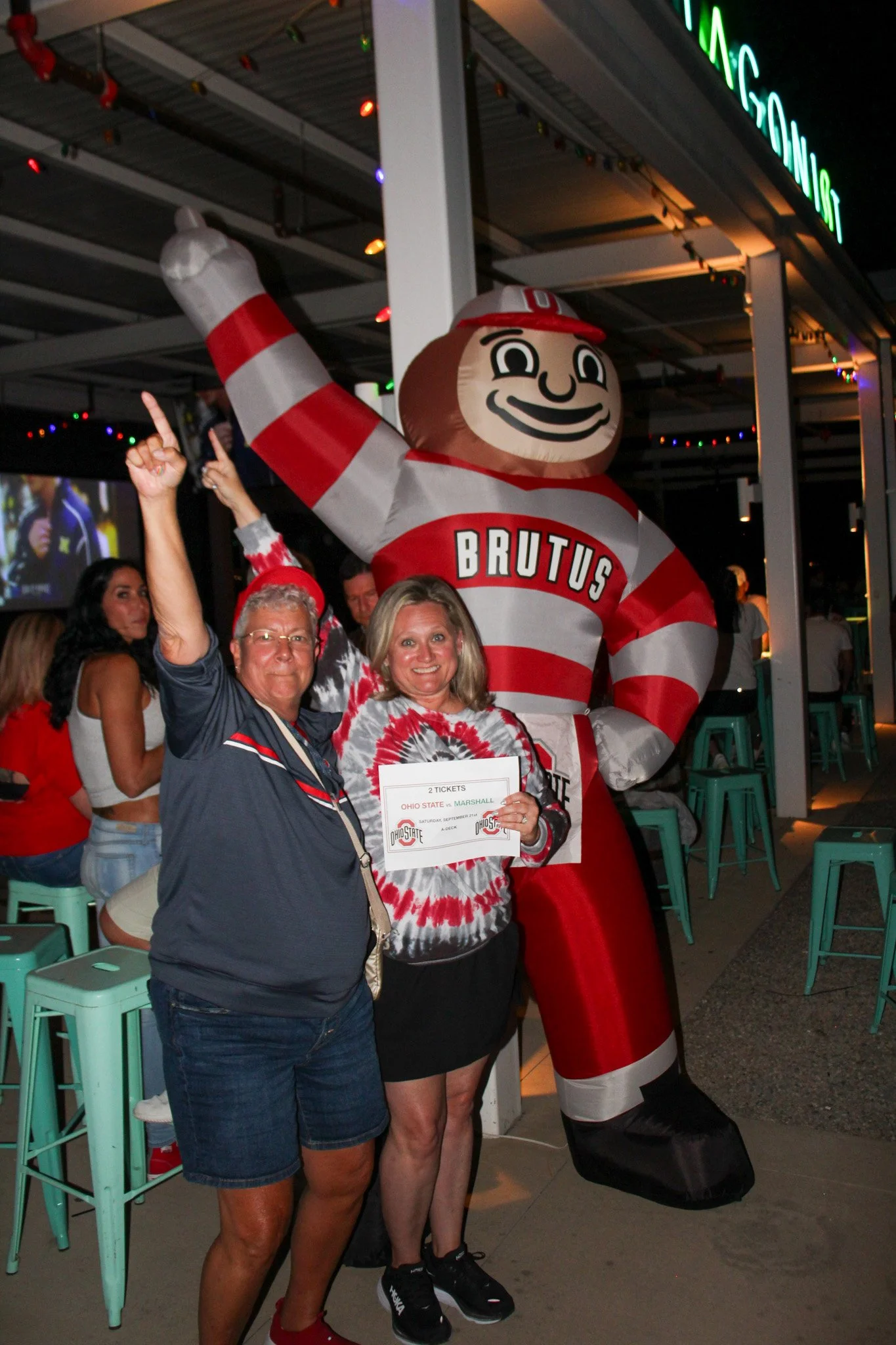 Two people celebrating with a large Brutus mascot, Ohio State University, holding tickets, at a sports event or party.