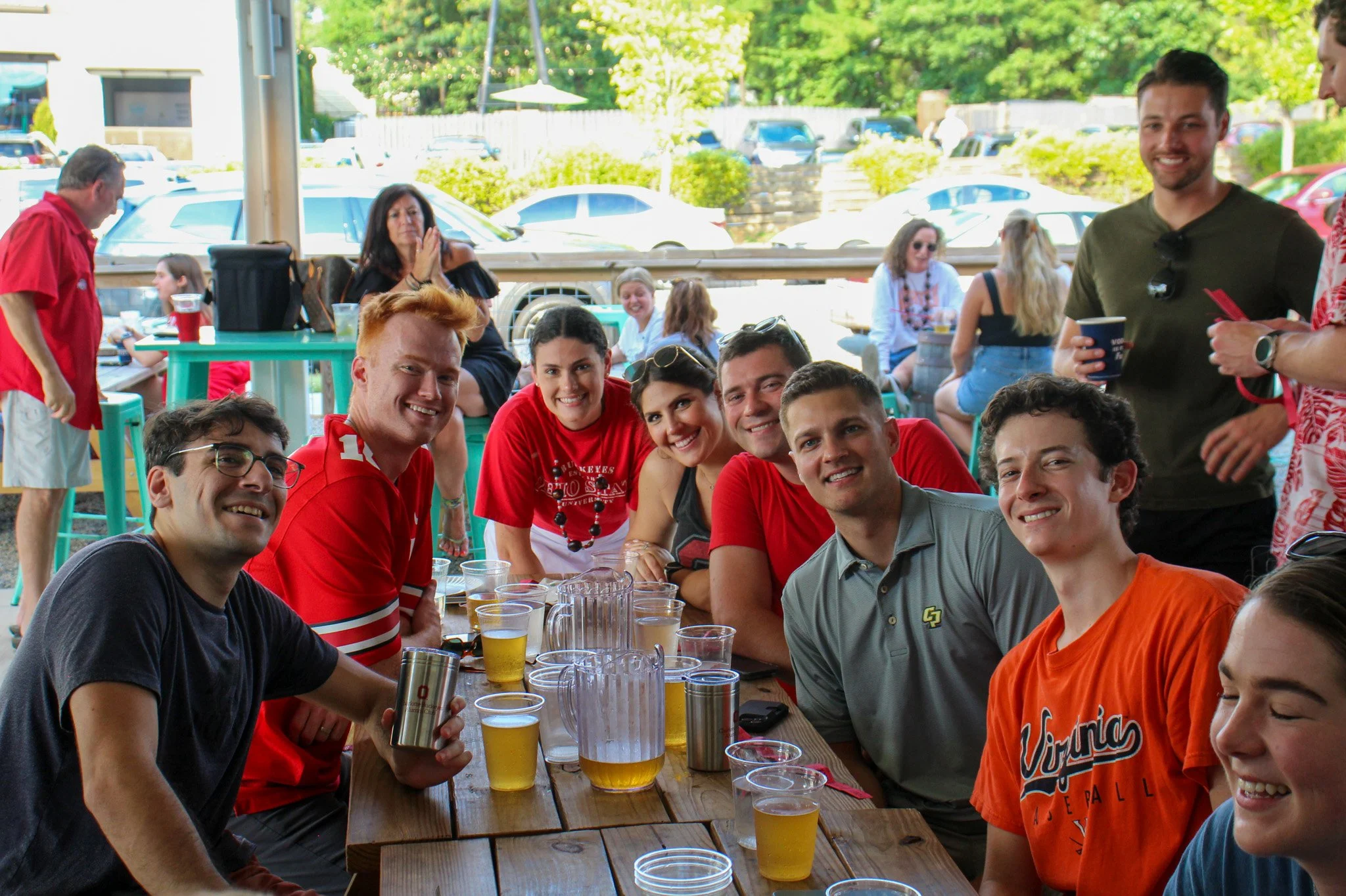 Group of friends smiling and sitting around a table with drinks at a backyard gathering on a sunny day.