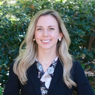 A woman with shoulder-length light brown hair, smiling, wearing pearl earrings and a black top outdoors with a blurred green background.
