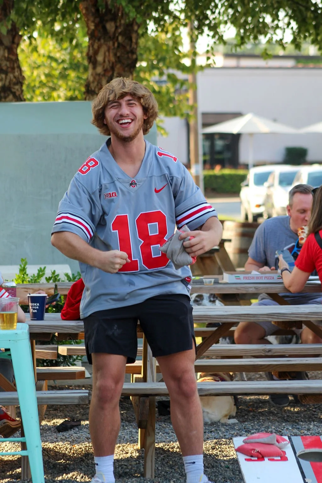 Young man smiling and cheering while wearing a gray Ohio State football jersey with red and white details and the number 18, outdoors at a gathering with picnic tables, other people, and trees in the background.