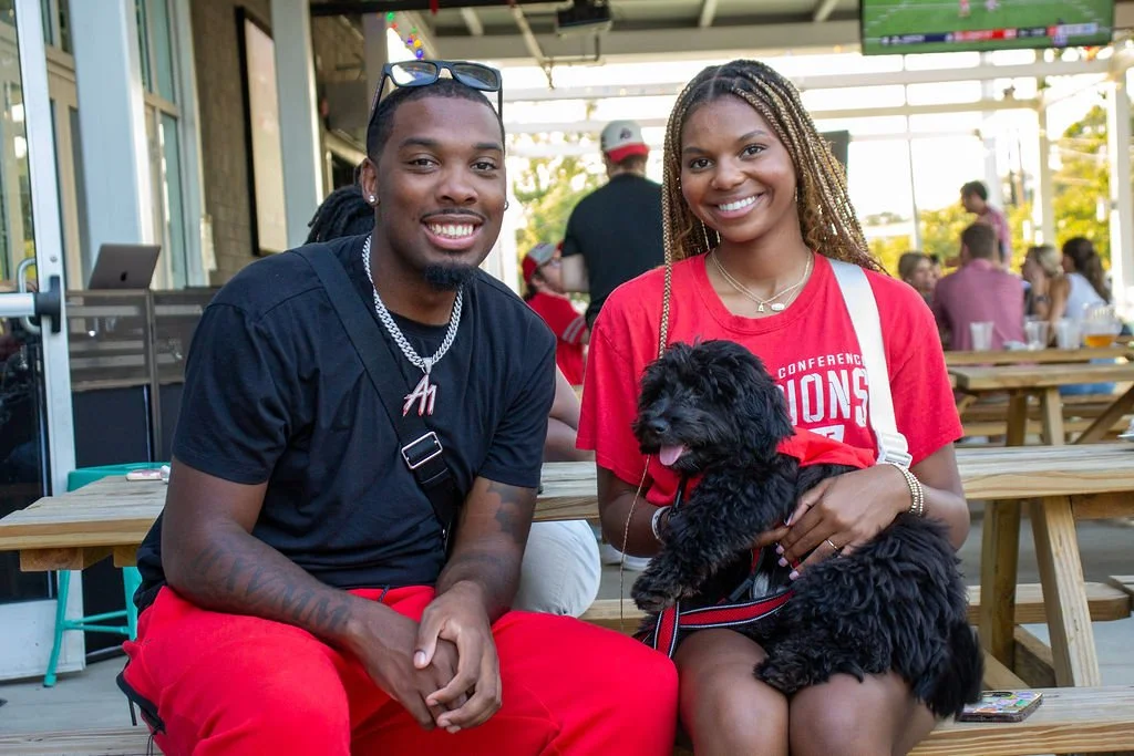 A smiling young man and woman sitting on a wooden picnic table outside, with the woman holding a small black dog. People are in the background at an a Charlotte OSU Game Watch