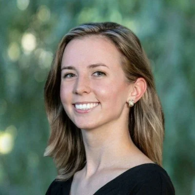 Portrait of a smiling woman with shoulder-length light brown hair, wearing a black top and pearl earrings, outdoors with blurred greenery in the background.