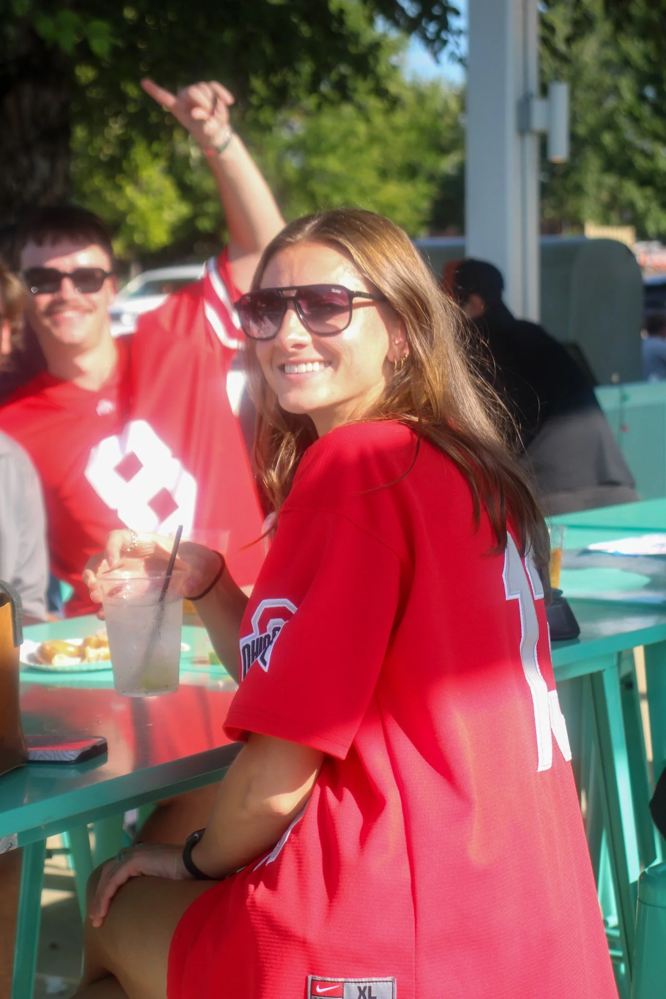 A woman wearing sunglasses and a red Ohio State sports jersey sitting at an outdoor table, smiling with a drink in hand, with people in red jerseys and trees in the background.