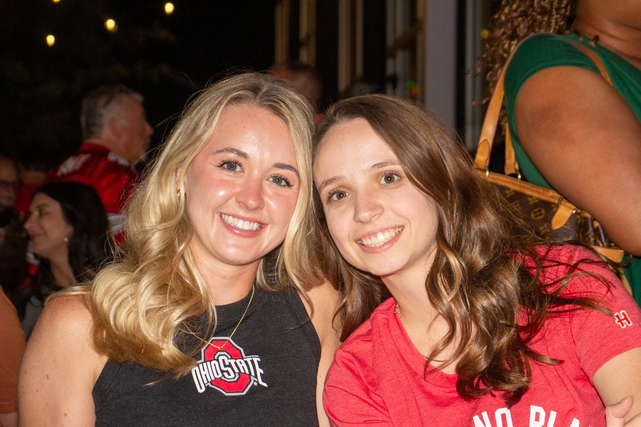 Two young women smiling closely together, both wearing red Ohio State shirts, at an outdoor event at night.