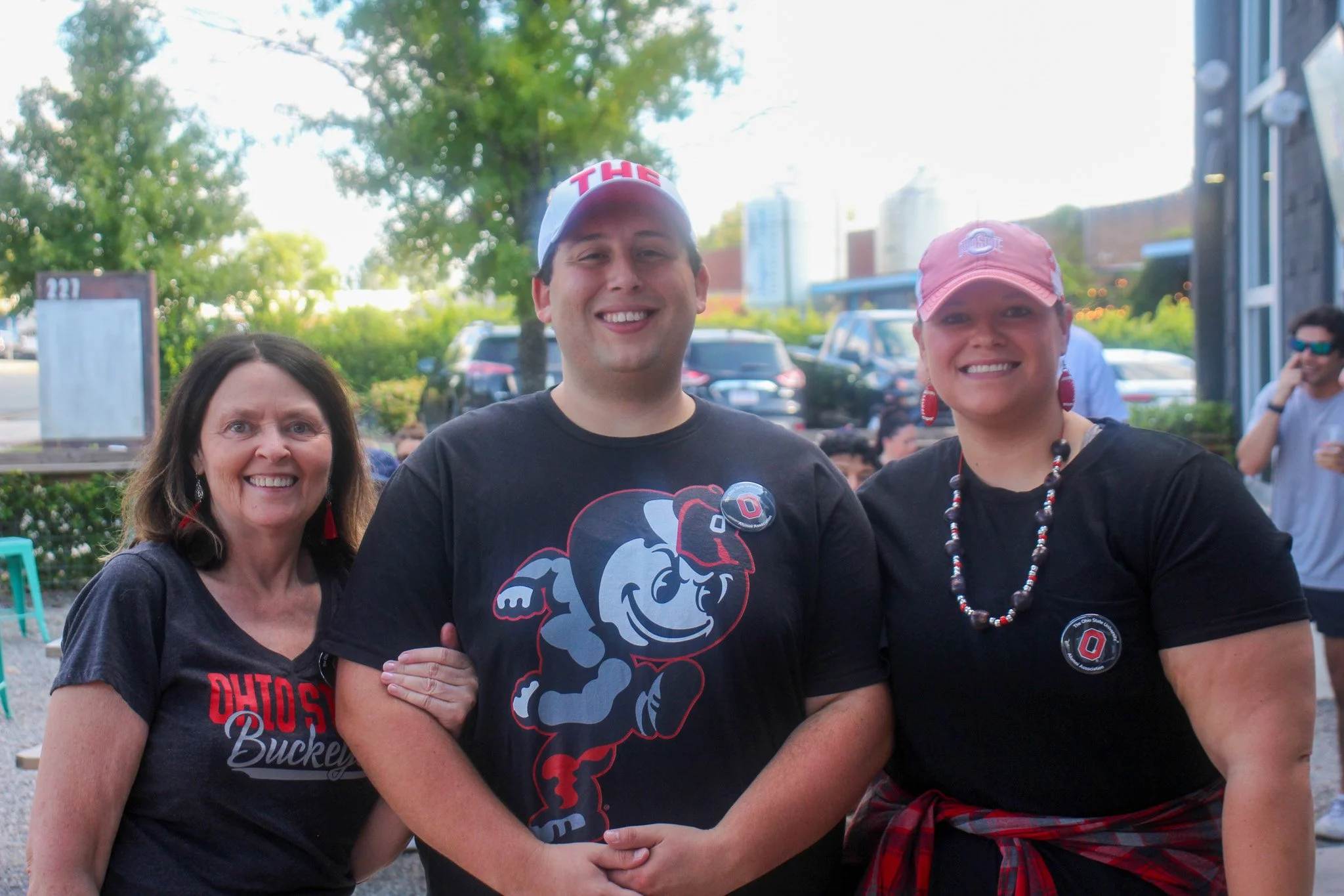 Group of three smiling people outdoors, wearing Ohio State Buckeyes-themed clothing and accessories, with trees and parked cars in the background.
