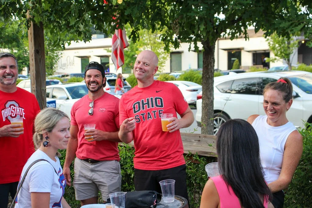 Group of people outdoors, wearing Ohio State shirts, standing around a table with drinks, smiling and talking.