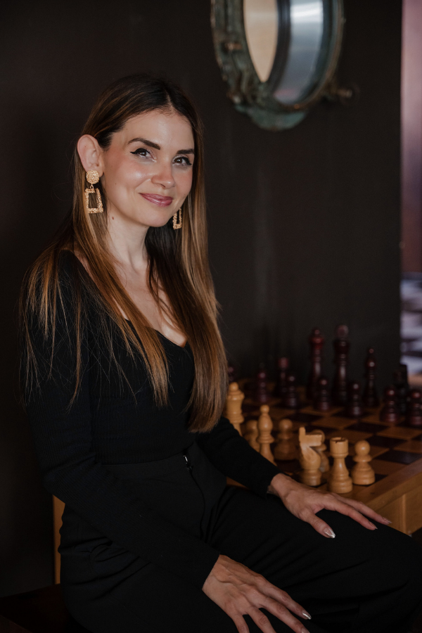 A woman with long brown hair wearing black clothing and gold earrings, sitting next to a chessboard with wooden chess pieces, in a dimly lit room.