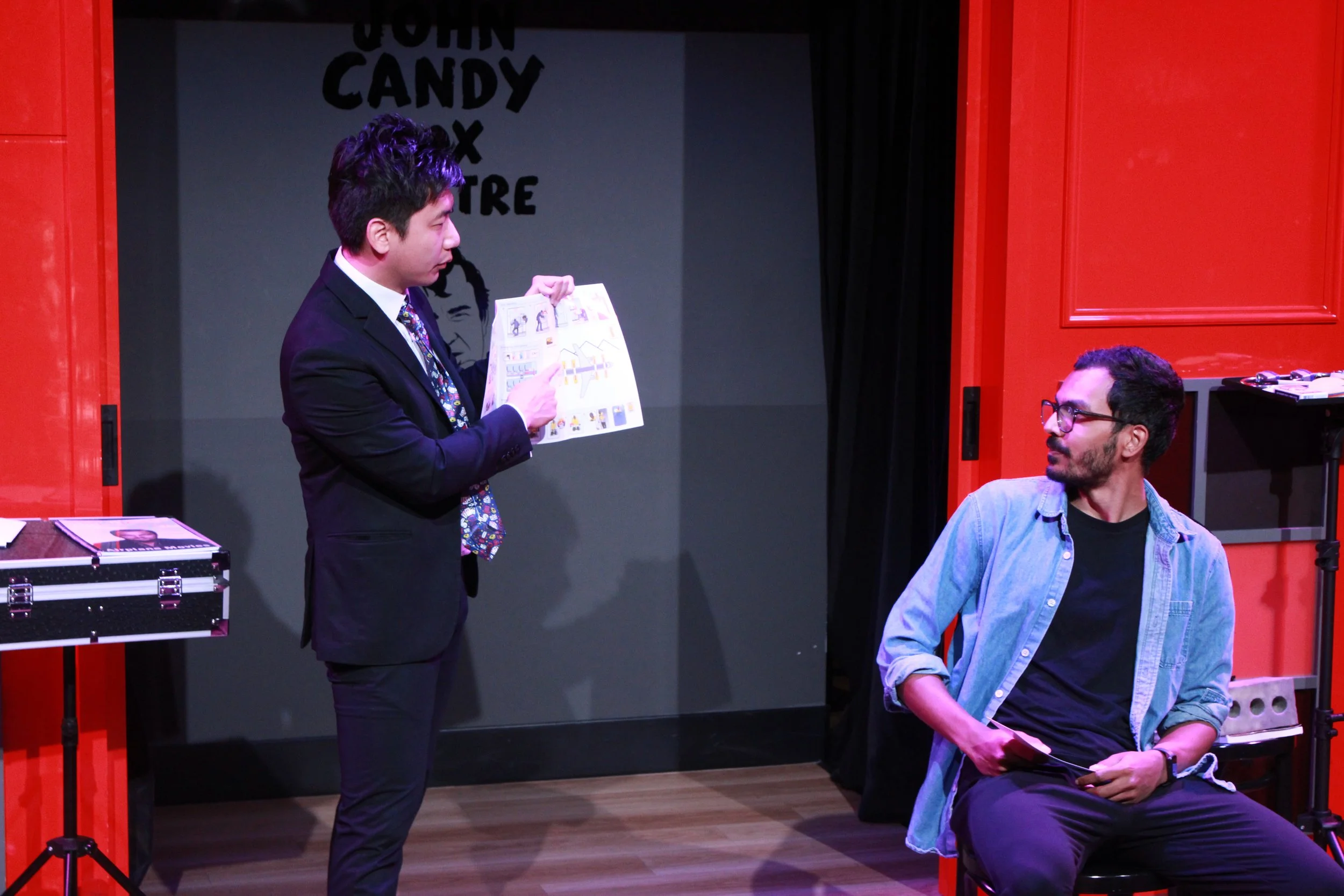 A man in a suit and a tie is standing and showing a comic book to a man in a gray shirt and glasses who is sitting on a stool. The scene takes place in a theater dressing room with red walls and black curtains in the background.