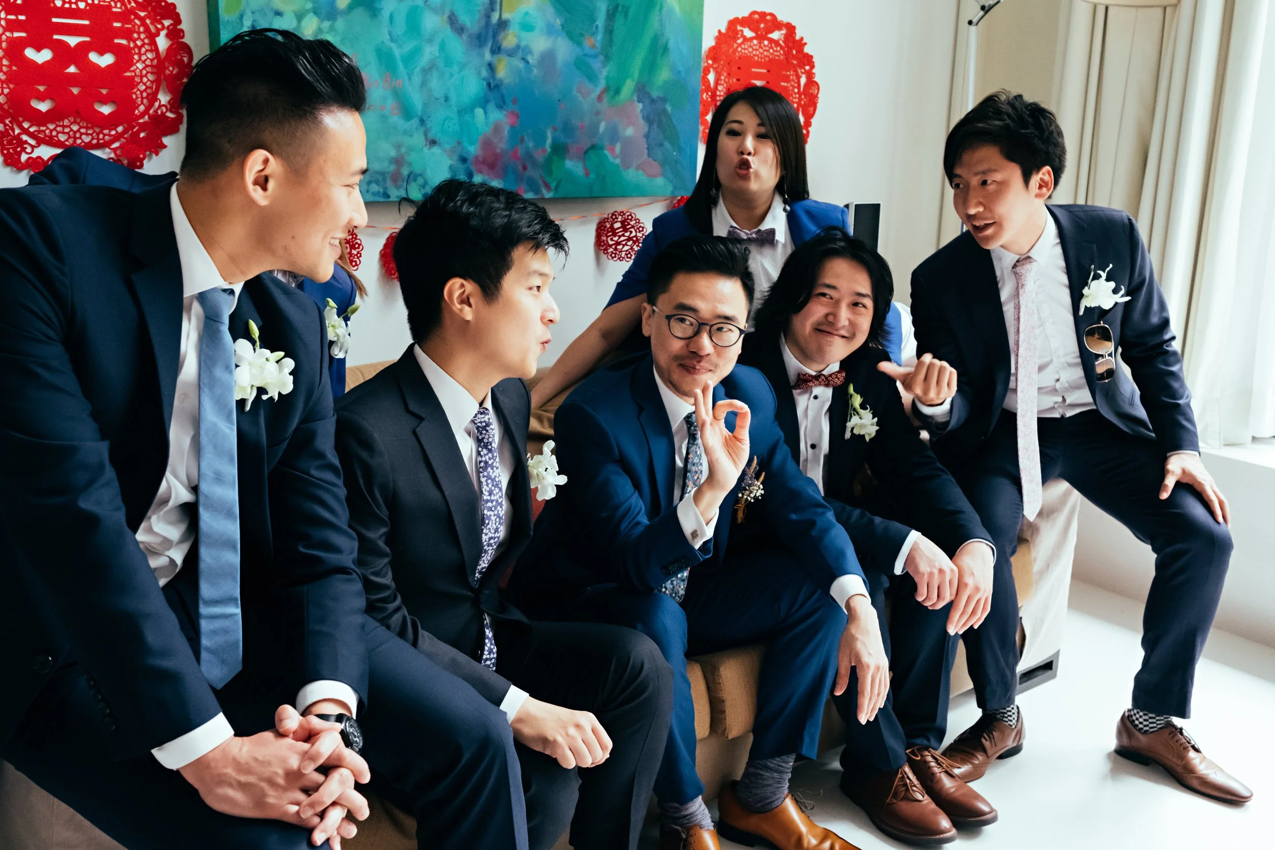 Group of young men and women dressed in formal suits and dresses, gathered indoors, enjoying lively conversation and photos during a celebration. Decorations include red paper cutouts and a colorful abstract painting in the background.