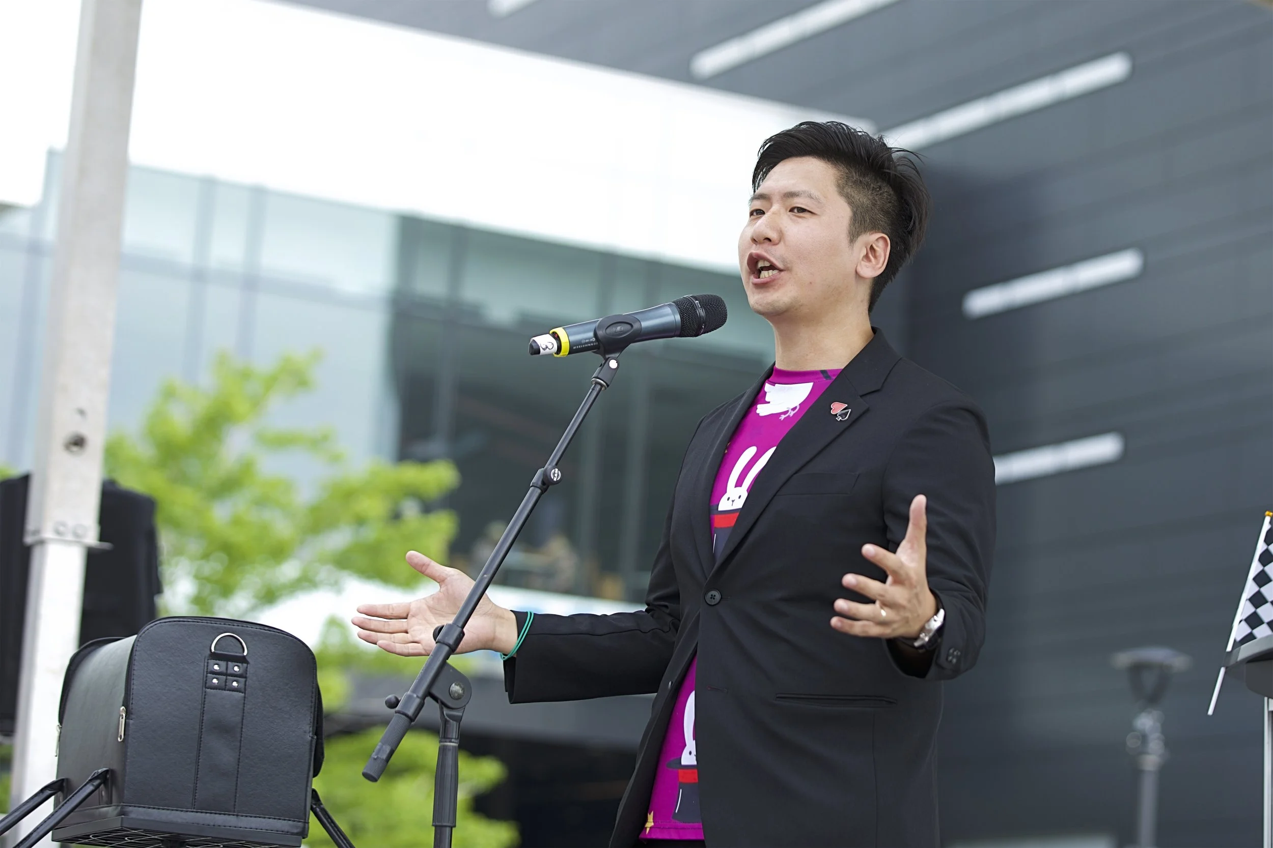 A man in a black blazer and pink shirt speaking into a microphone at an outdoor event. He is using hand gestures to emphasize his speech, with a black bag on a chair nearby and modern buildings in the background.