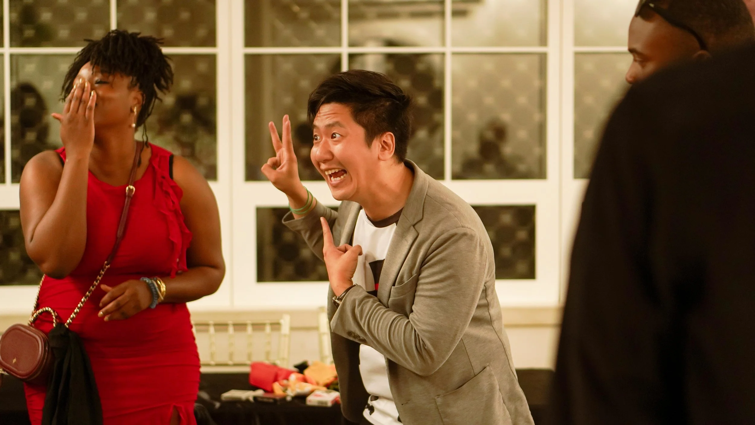 A group of three people interacting at a social gathering indoors. A woman in a red dress covering her face, a man in a gray blazer making a funny gesture with his hand, and another man partially visible on the right side of the image.