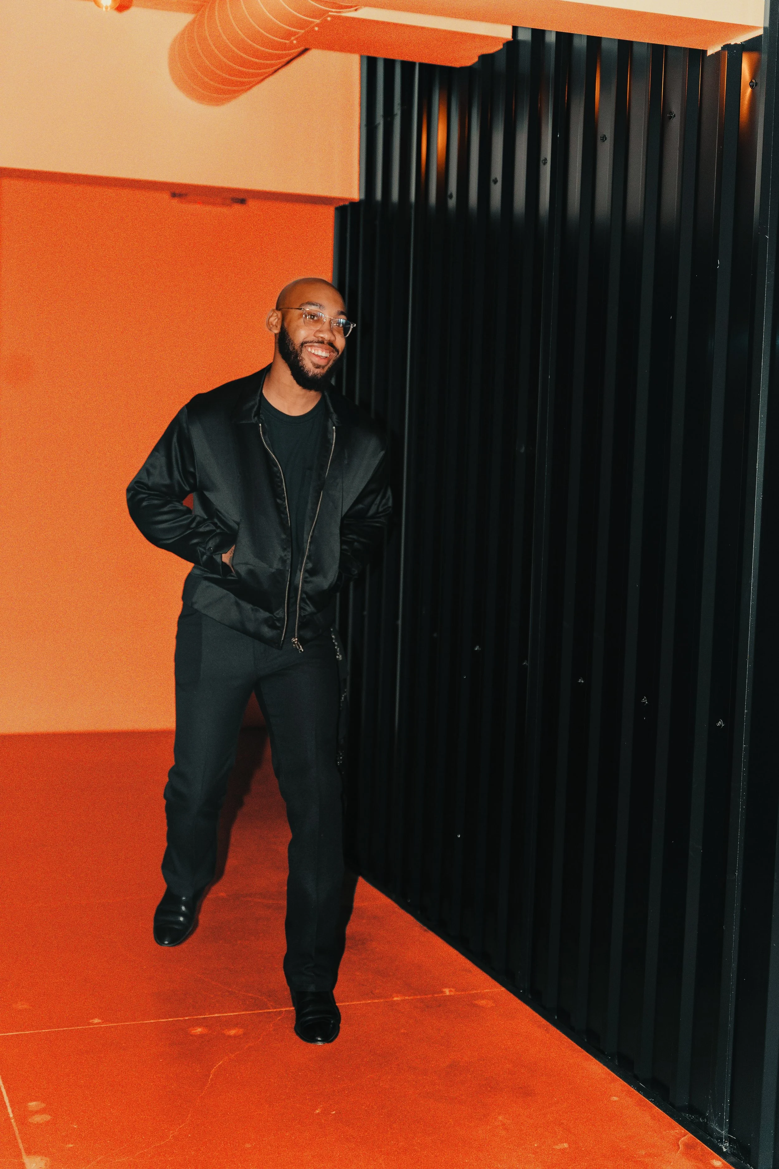 A man with a beard, glasses, and a bald head smiles while walking indoors near a black metal wall. He is wearing a black satin bomber jacket, black pants, and black shoes.