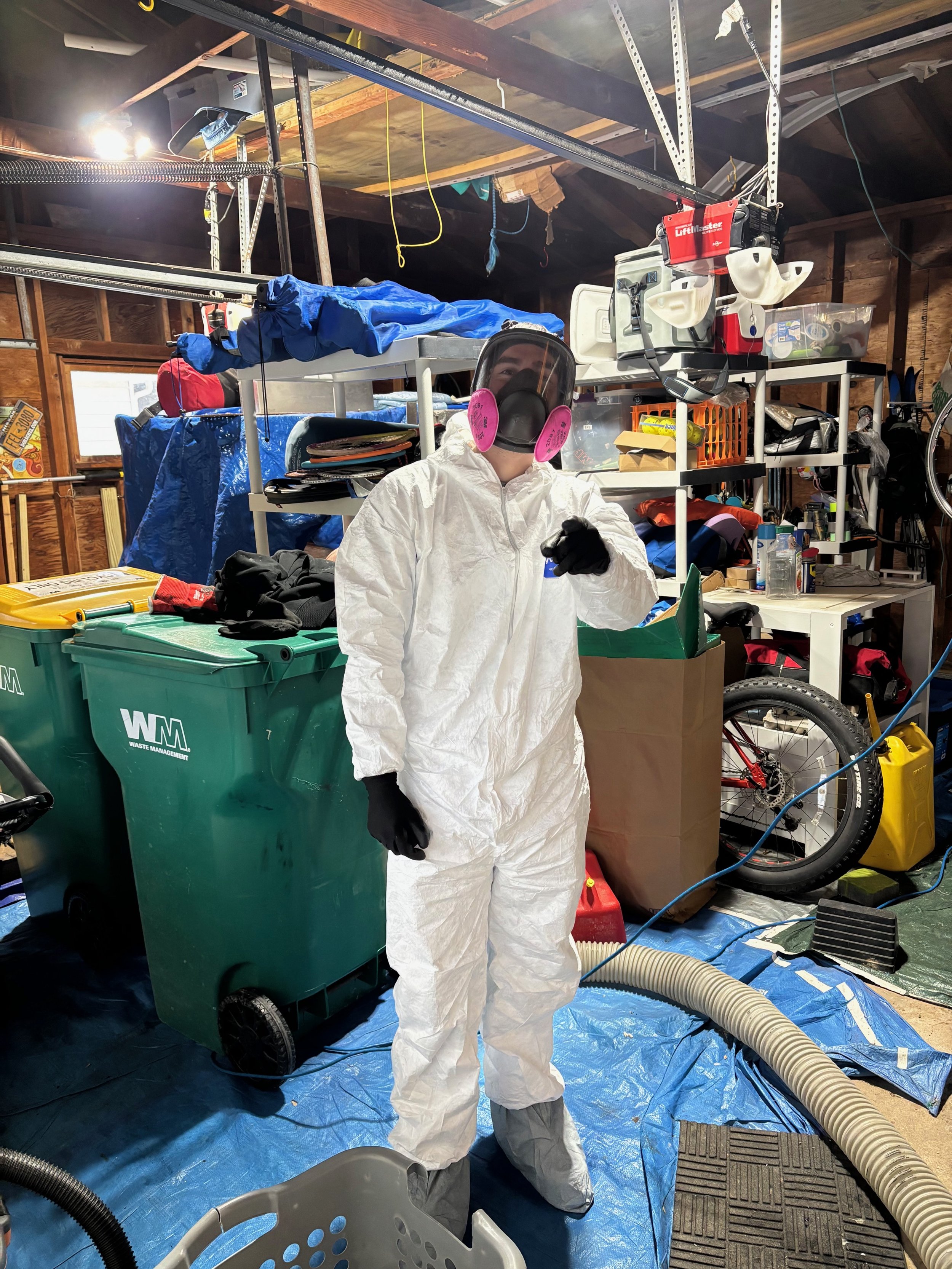 Person dressed in a white hazmat suit with black gloves and a face mask with pink filters, standing in a cluttered garage or workshop with various tools, bikes, and storage boxes around.