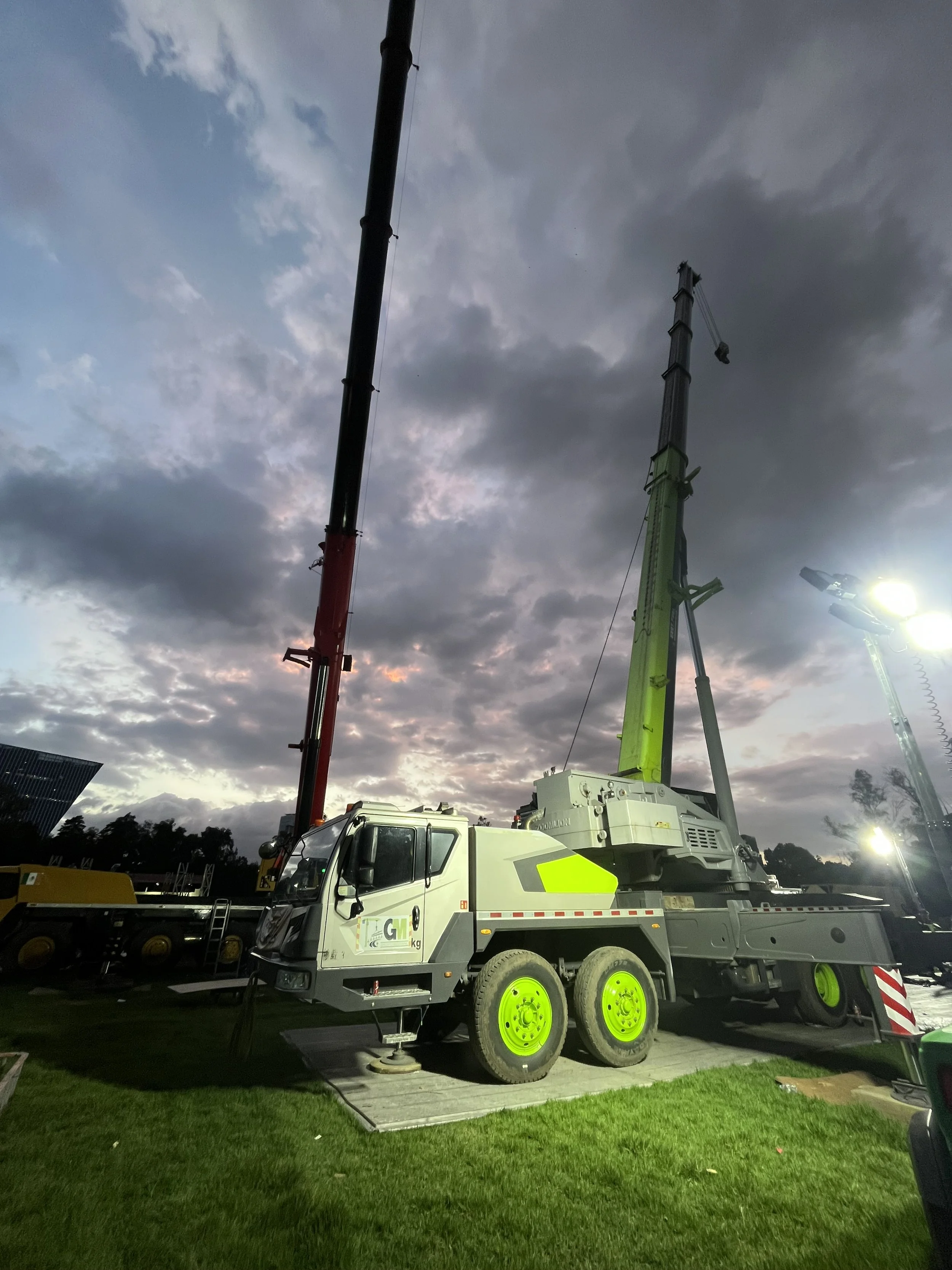 A mobile crane with bright yellow wheels and a white body, working outdoors at dusk with a cloudy sky overhead. The crane's arm is extended upward and a crane hook hangs at the end.