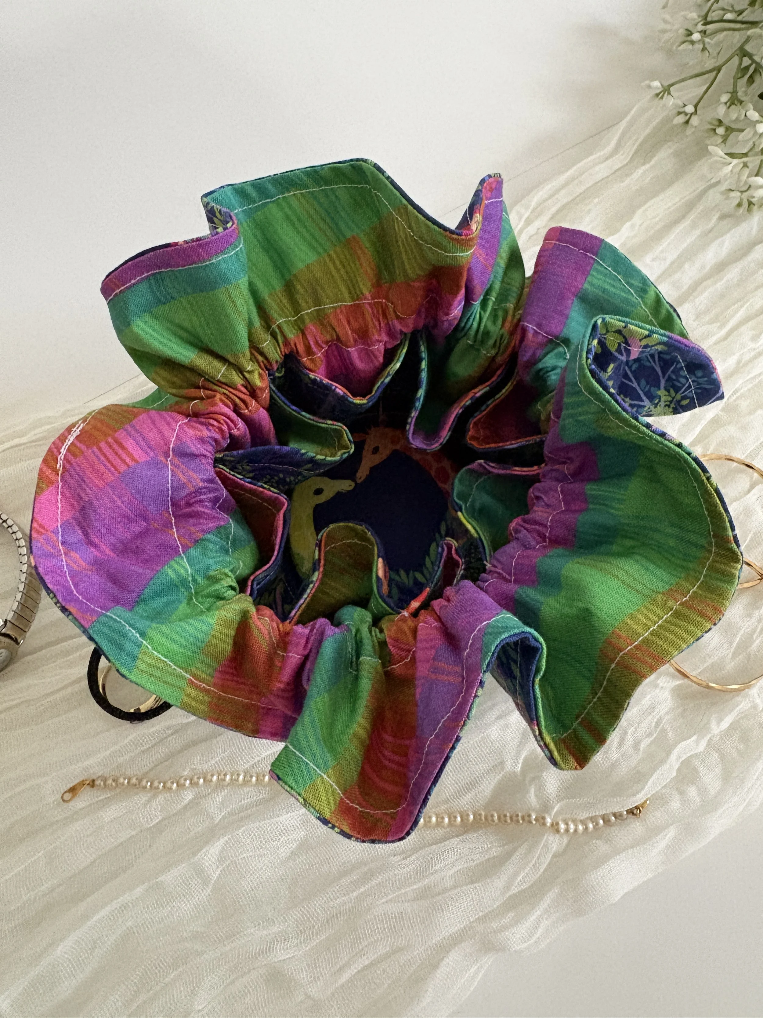 Colorful fabric with multiple shades of pink, green, purple, and blue, arranged in a ruffled circular shape on a textured white surface, with a string of pearls and a gold zipper nearby.