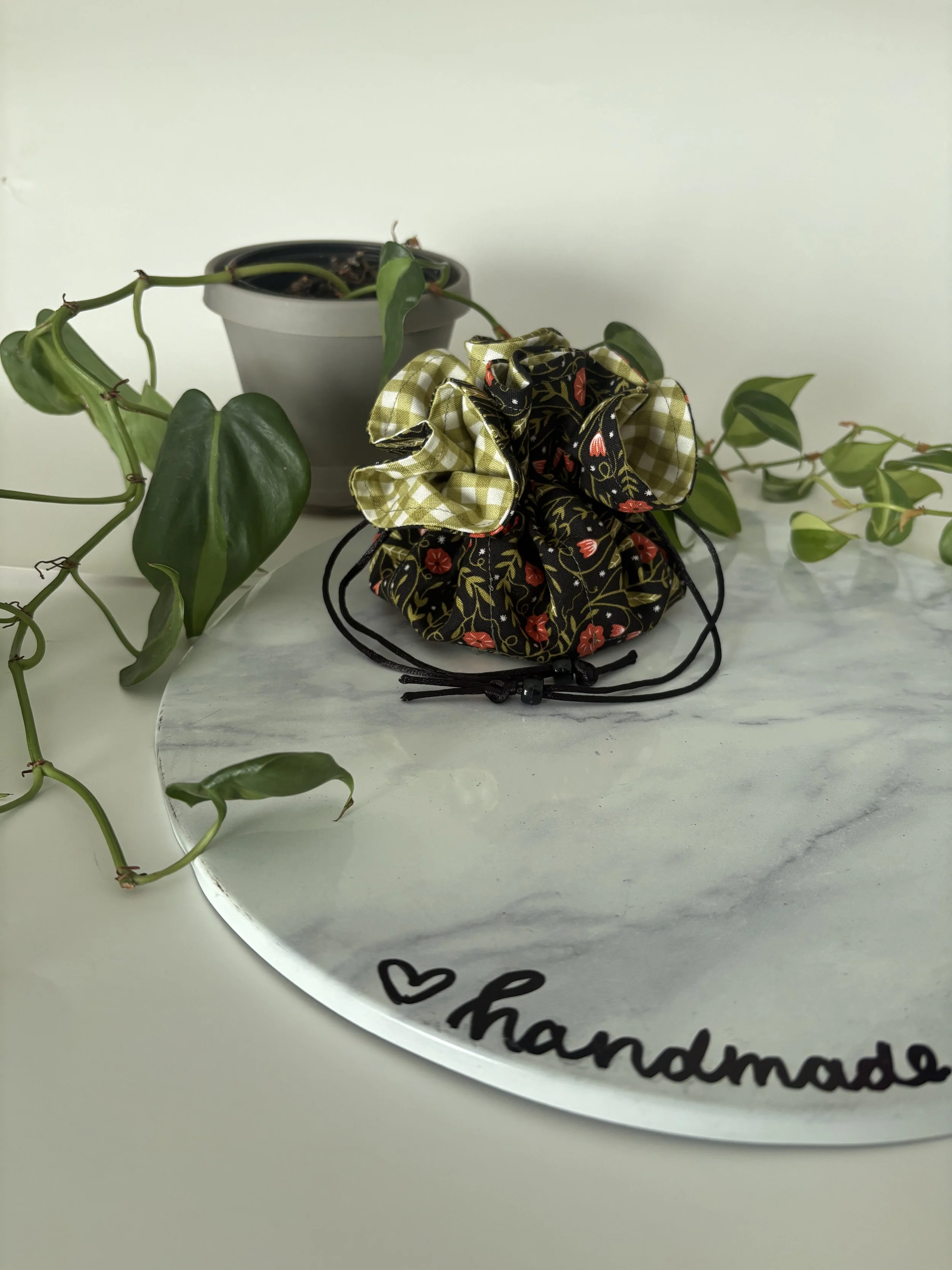 A decorative cloth gift bag with red, black, and green botanical patterns, resting on a white circular surface with the partial word "Handmade" written in black and a small heart symbol.