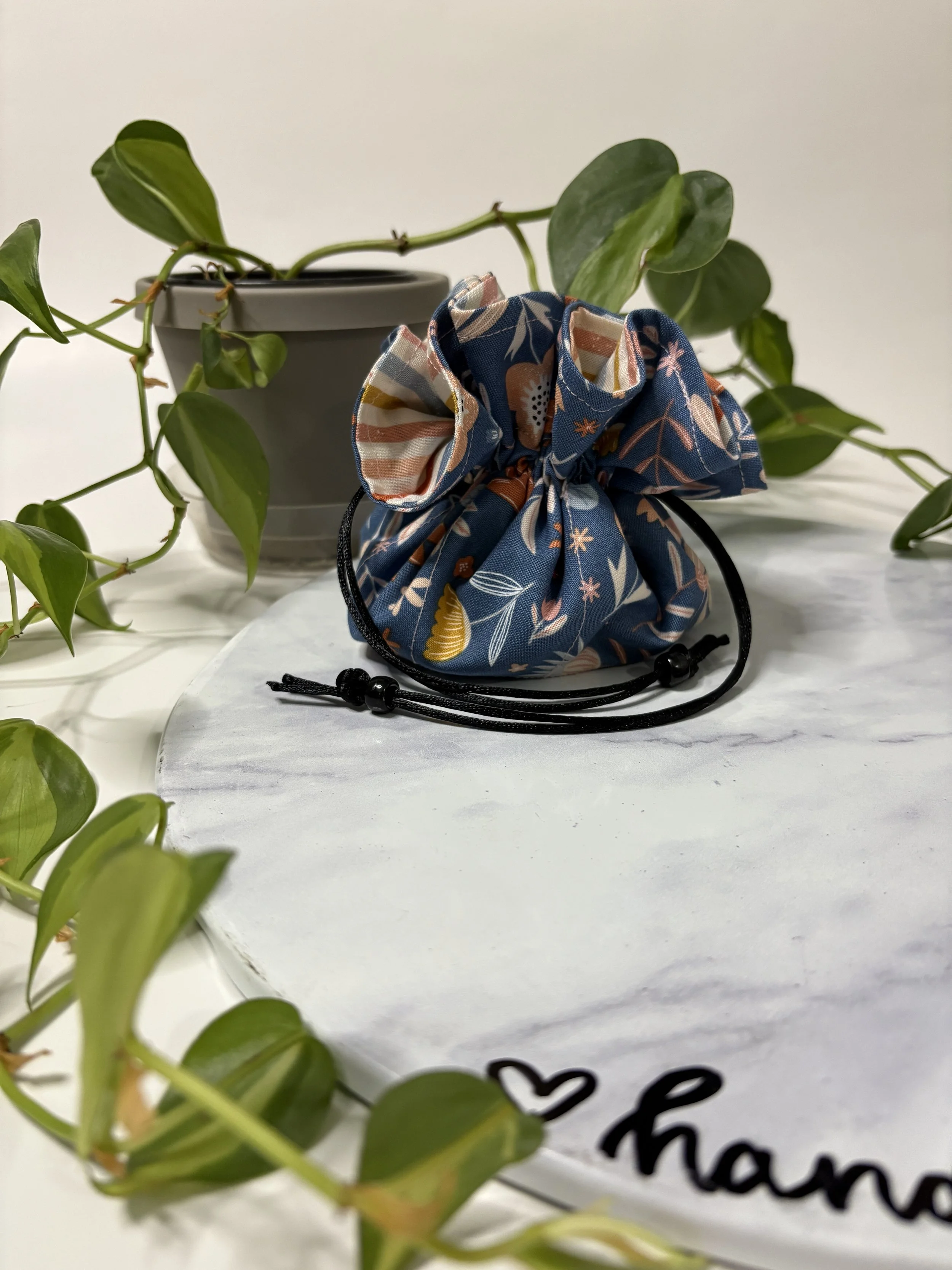A potted green plant with trailing vines, next to a fabric drawstring bag with a floral pattern, on a white surface with a partially visible handwritten message.