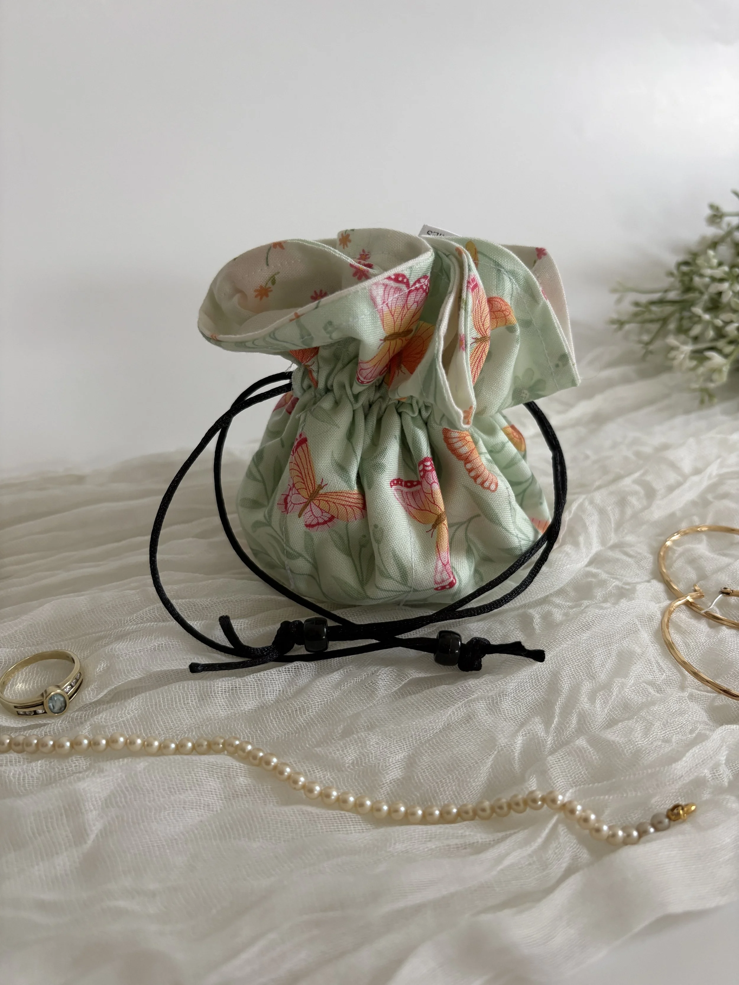 A floral fabric pouch with drawstrings, placed on white fabric, surrounded by jewelry including a pearl necklace, a gold ring, a gold bracelet, and a gold ring with a gemstone, with a small sprig of white flowers on the right.