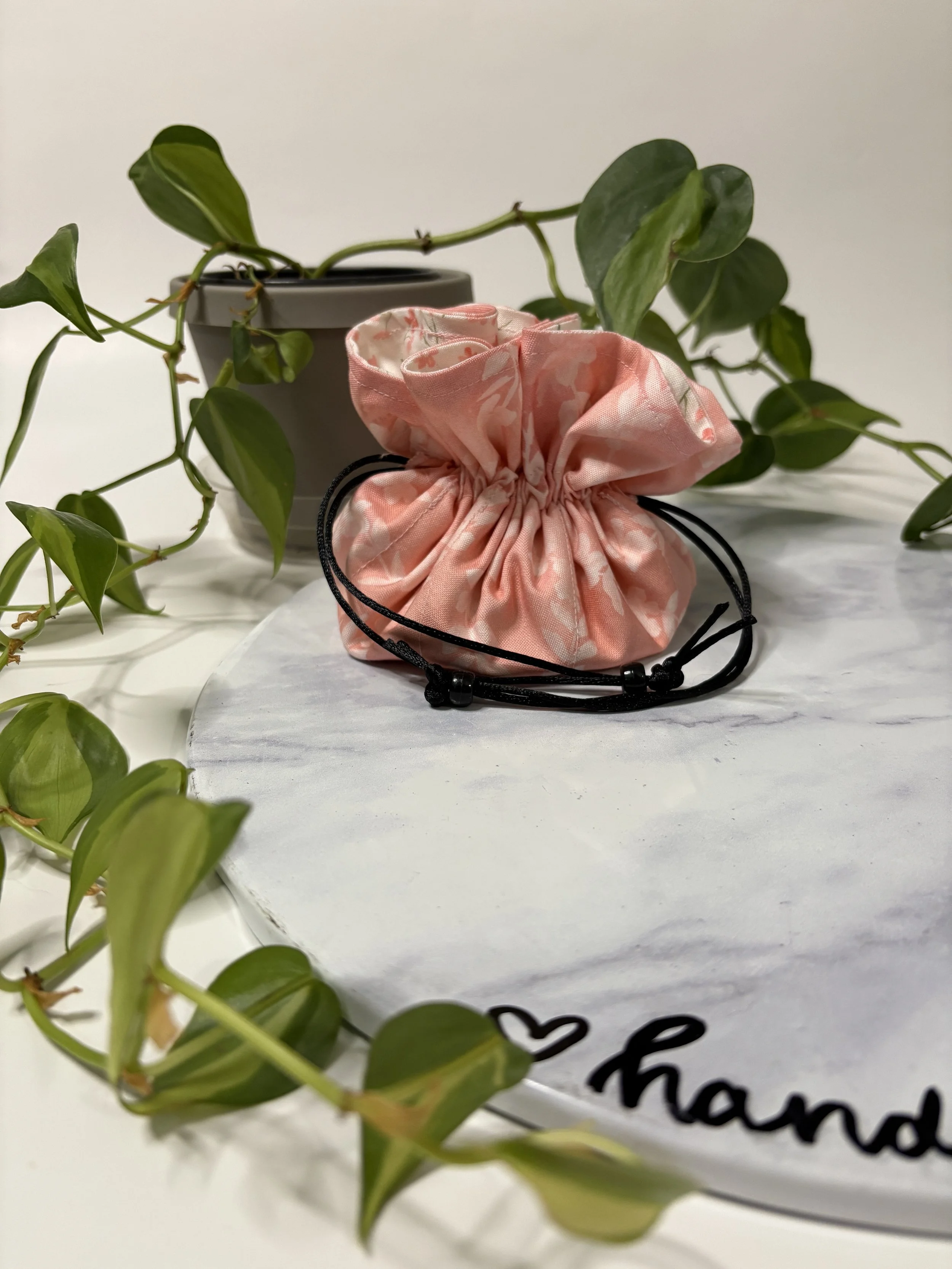 A pink, floral-patterned fabric pouch with a black drawstring, placed on a white surface in front of a potted plant with green leaves.