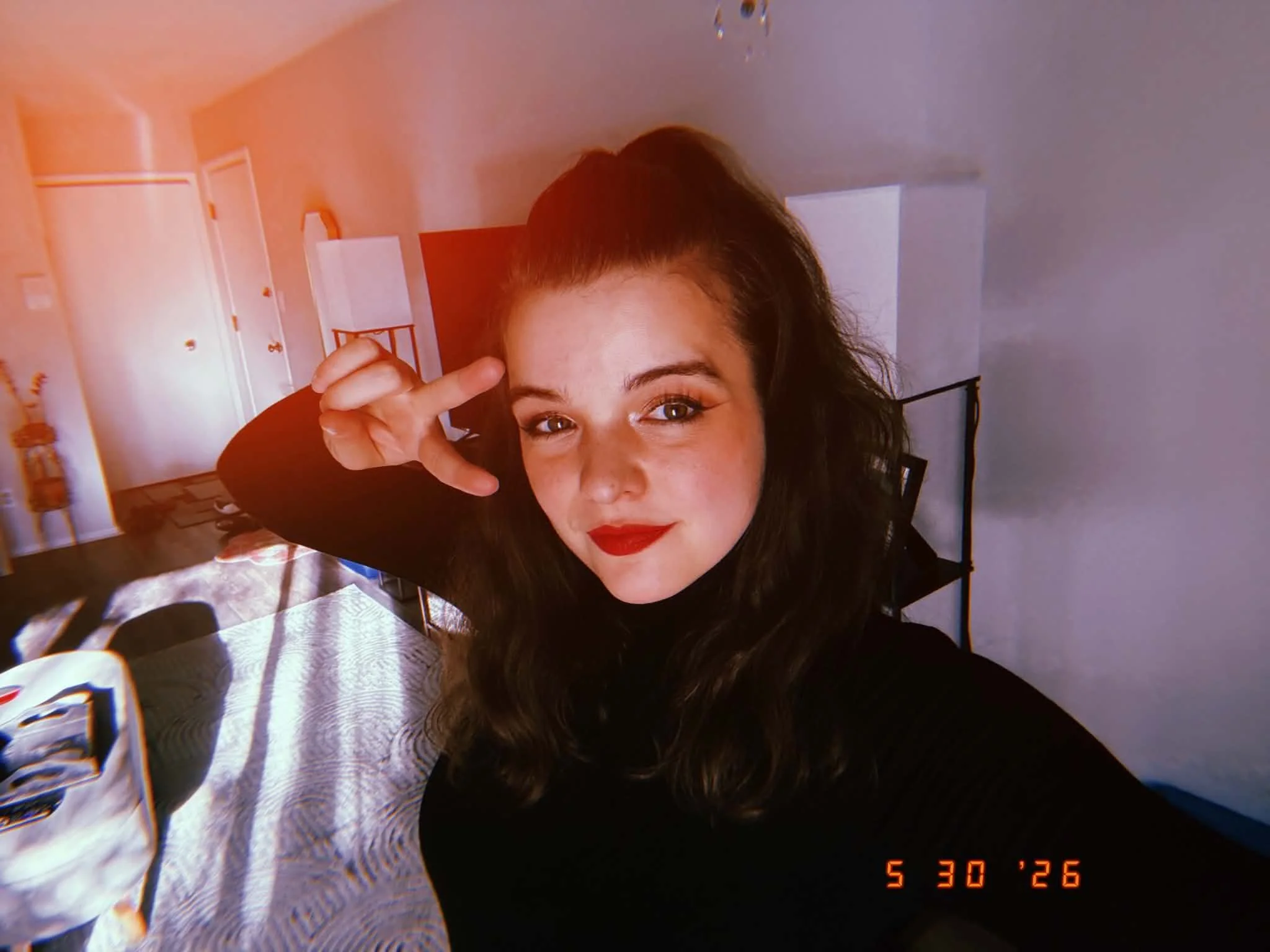 A woman with long dark hair and red lipstick taking a selfie inside a house, with a bedroom in the background.
