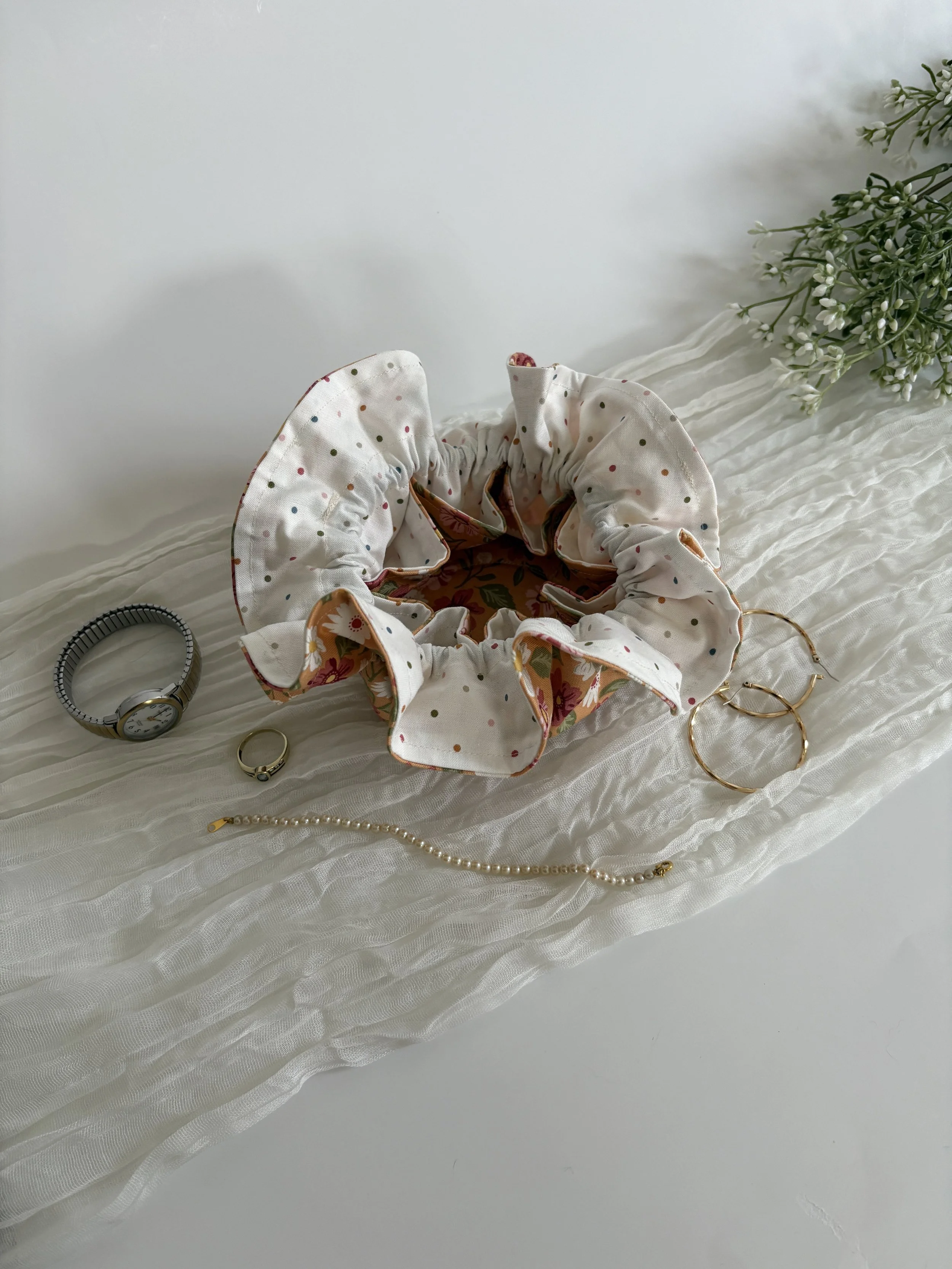 A collection of jewelry and a hair scrunchie arranged on a white, textured fabric surface, with some flowers in the background.