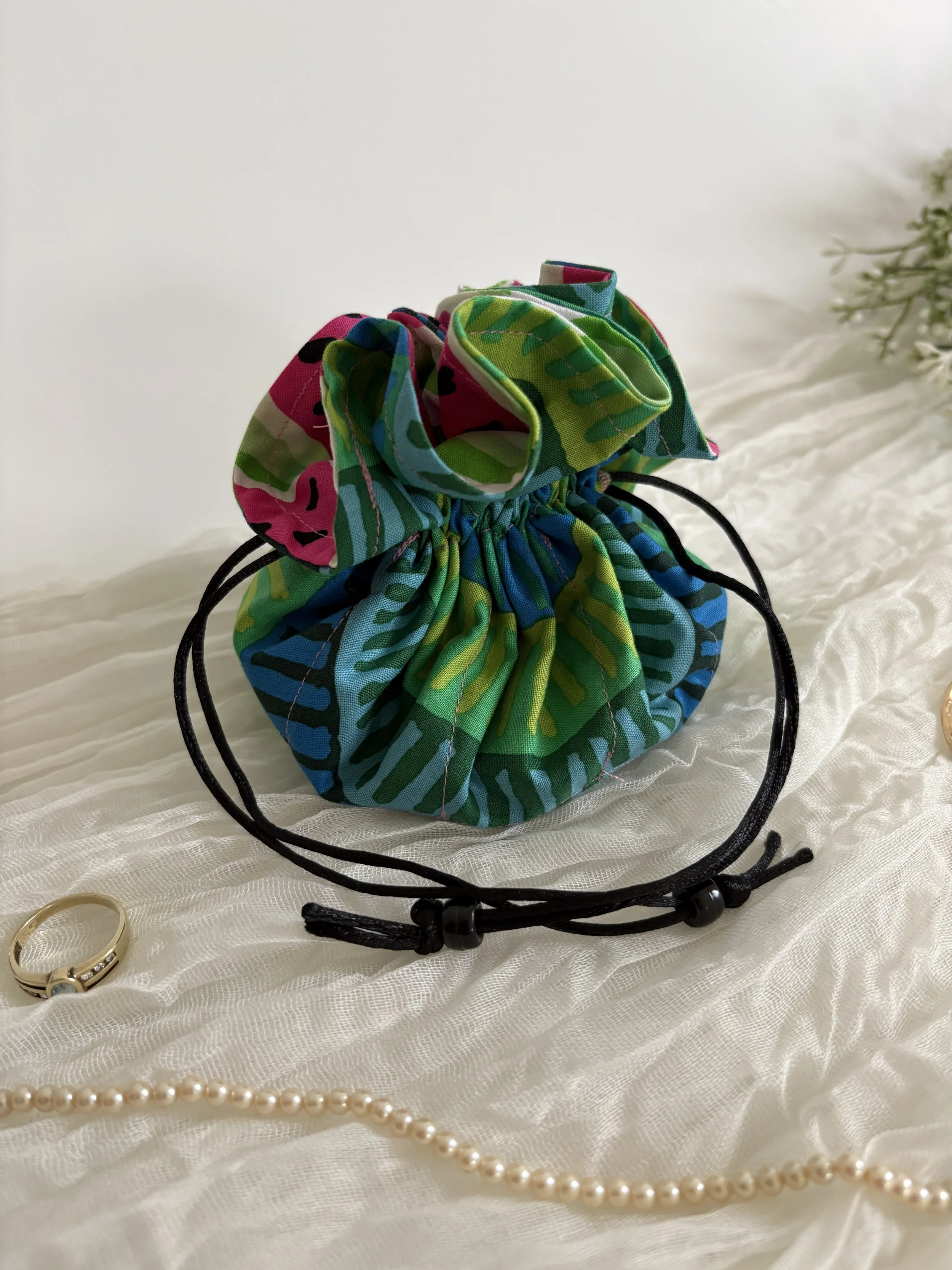 A fabric drawstring bag with a colorful, abstract pattern in green, blue, pink, and yellow, sitting on white fabric with jewelry items including a pearl necklace, gold rings, and a black leather bracelet intertwined nearby.